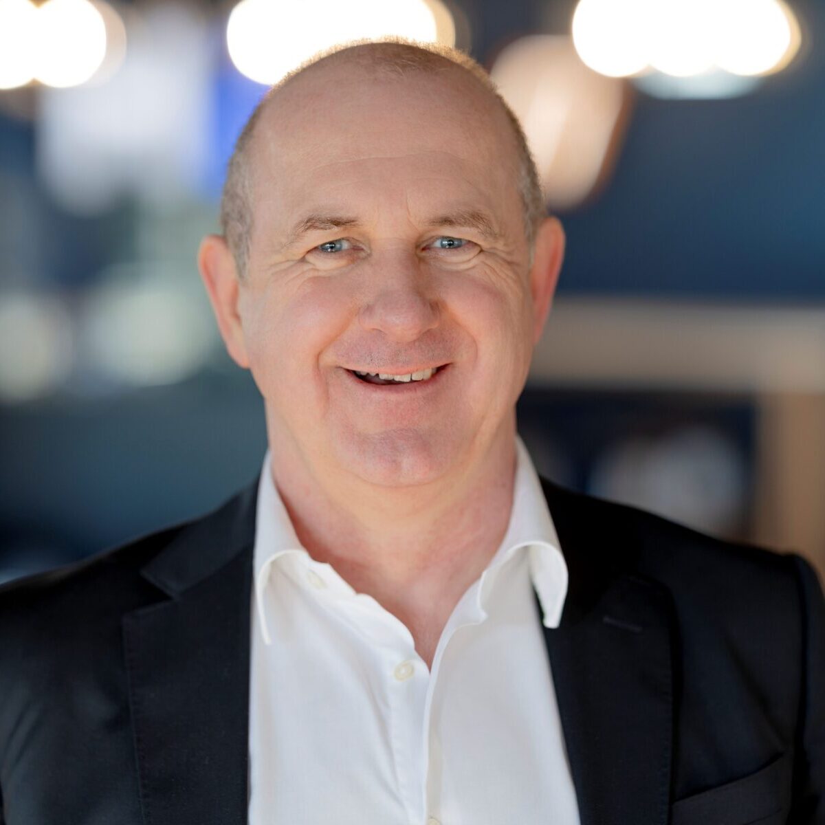 Ein Mann mittleren Alters mit kurzen Haaren lächelt in die Kamera. Er trägt ein weißes Hemd und einen dunklen Blazer. Der Hintergrund ist sanft verschwommen, mit warmen Lichtern und Blautönen.