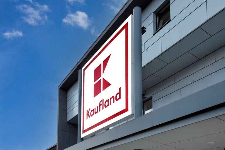 An der Fassade eines modernen Gebäudes ist ein großes, quadratisches Kaufland-Schild mit rot-weißem Logo angebracht. Im Hintergrund ist der Himmel klar und blau.