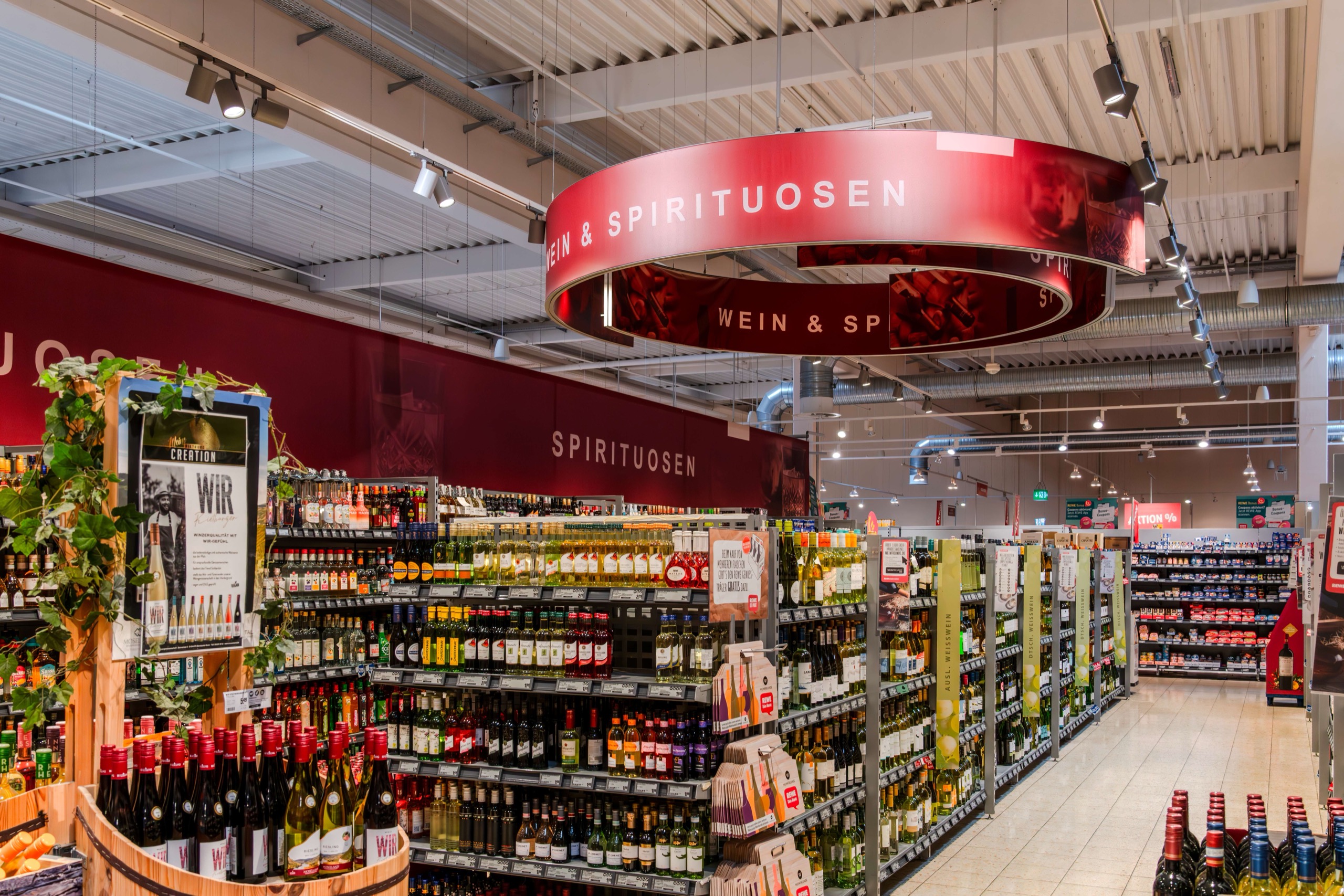 Ein hell erleuchteter Supermarktgang mit Regalen voller Wein und Spirituosen. Ein großes, rotes, rundes Schild mit der Aufschrift WEIN & SPIRITUOSEN steht über dem Regal, die Flaschen sind fein säuberlich aufgereiht, auf der linken Seite befinden sich einige Werbeaufsteller.