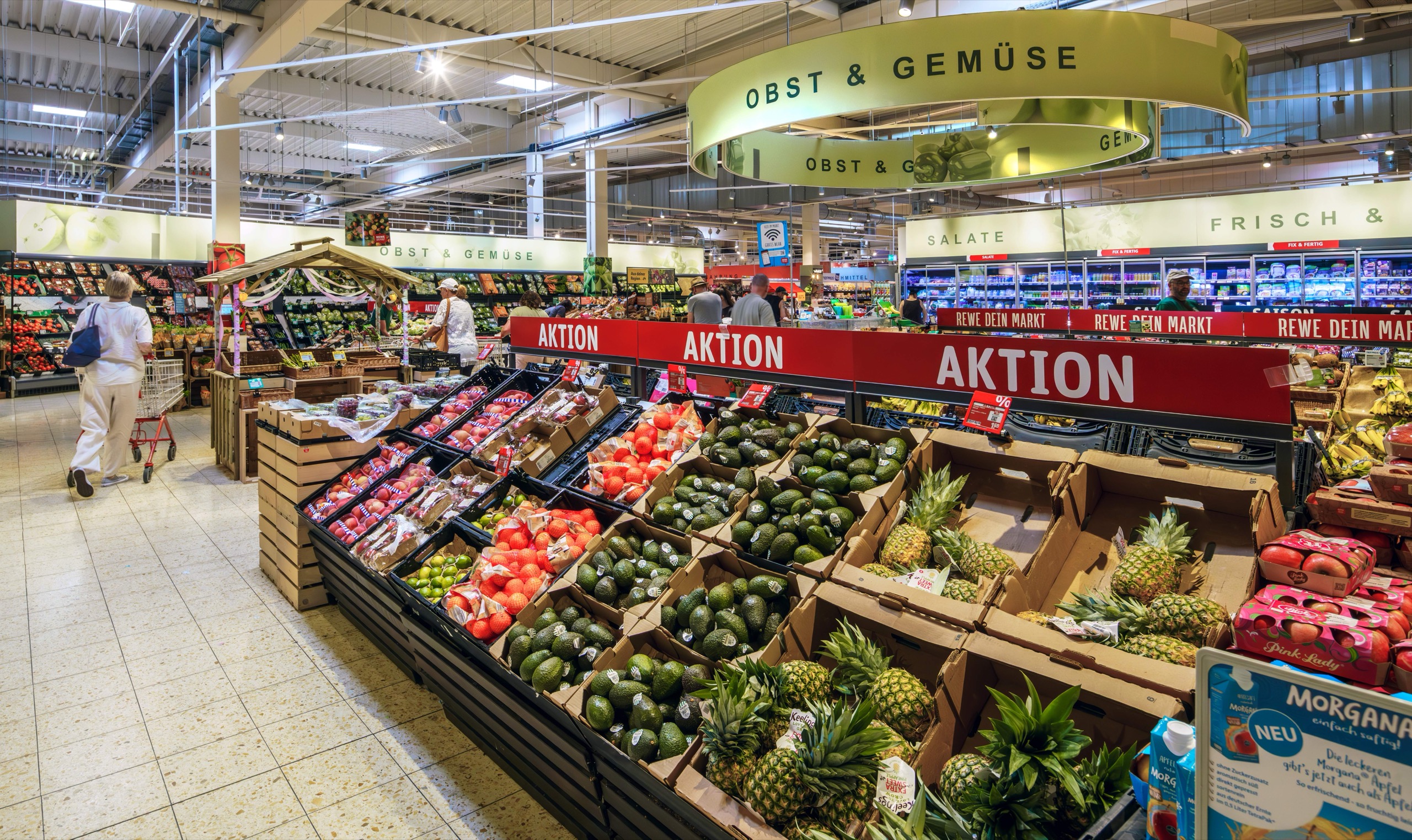 Ein hell erleuchteter Supermarkt mit Obst- und Gemüseauslagen, darunter Avocados, Ananas und Tomaten. Ein Kunde mit einem Einkaufswagen geht unter Schildern mit der Aufschrift Obst & Gemüse den Gang entlang.
