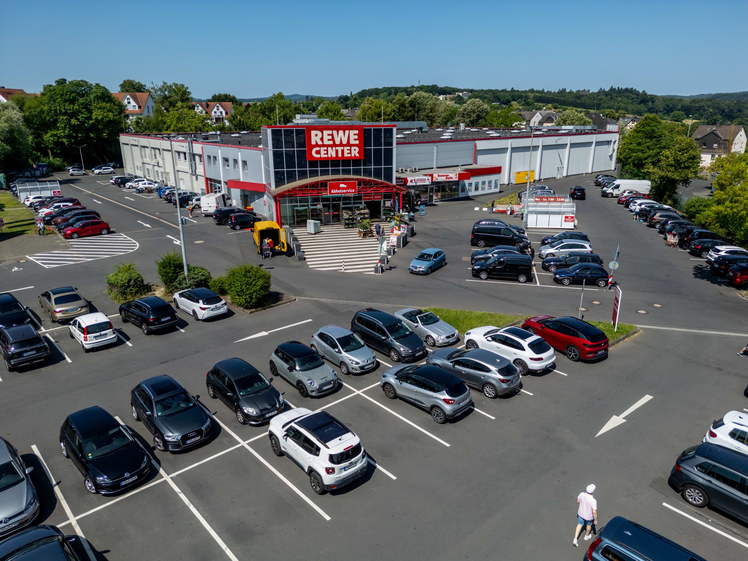 Luftaufnahme eines REWE-Center-Supermarkts mit einem überfüllten Parkplatz und mehreren geparkten Autos. Ein paar Leute gehen in der Nähe des Eingangs und auf dem Parkplatz spazieren, im Hintergrund sind Bäume und Häuser zu sehen.