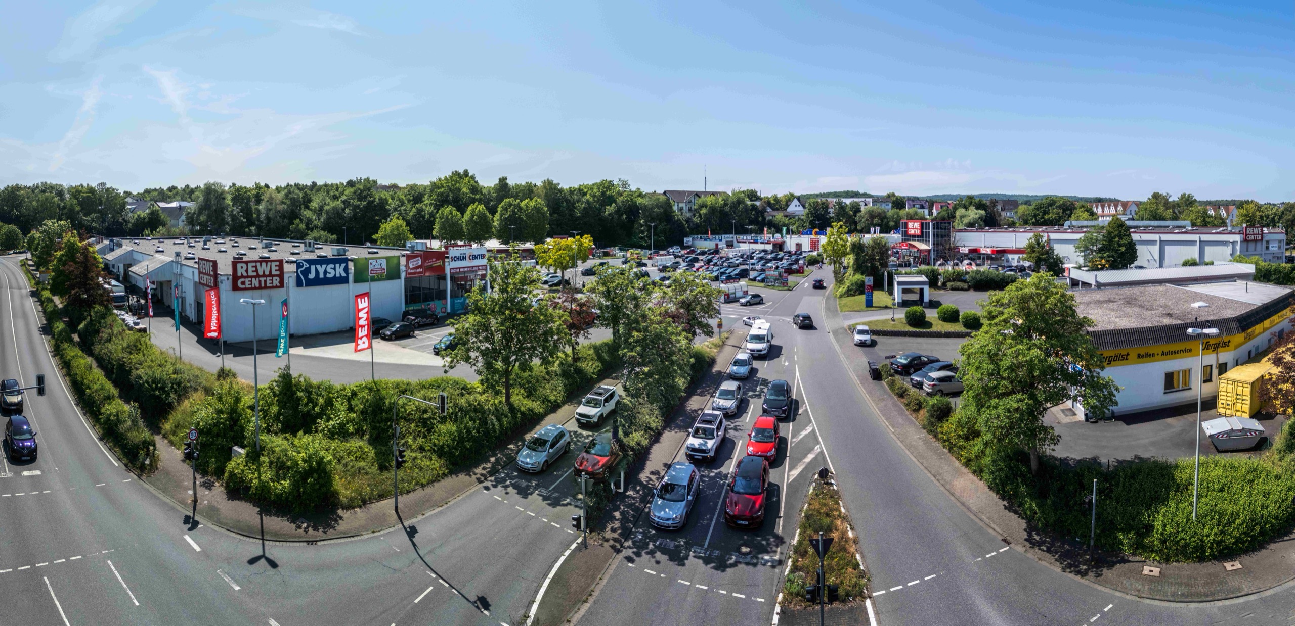 Luftaufnahme eines Einkaufszentrums mit einem großen Parkplatz, mehreren parkenden und fahrenden Autos und mehreren Geschäften, darunter REWE Center und JYSK, an einem sonnigen Tag. Bäume und Straßen umgeben das Gebiet.