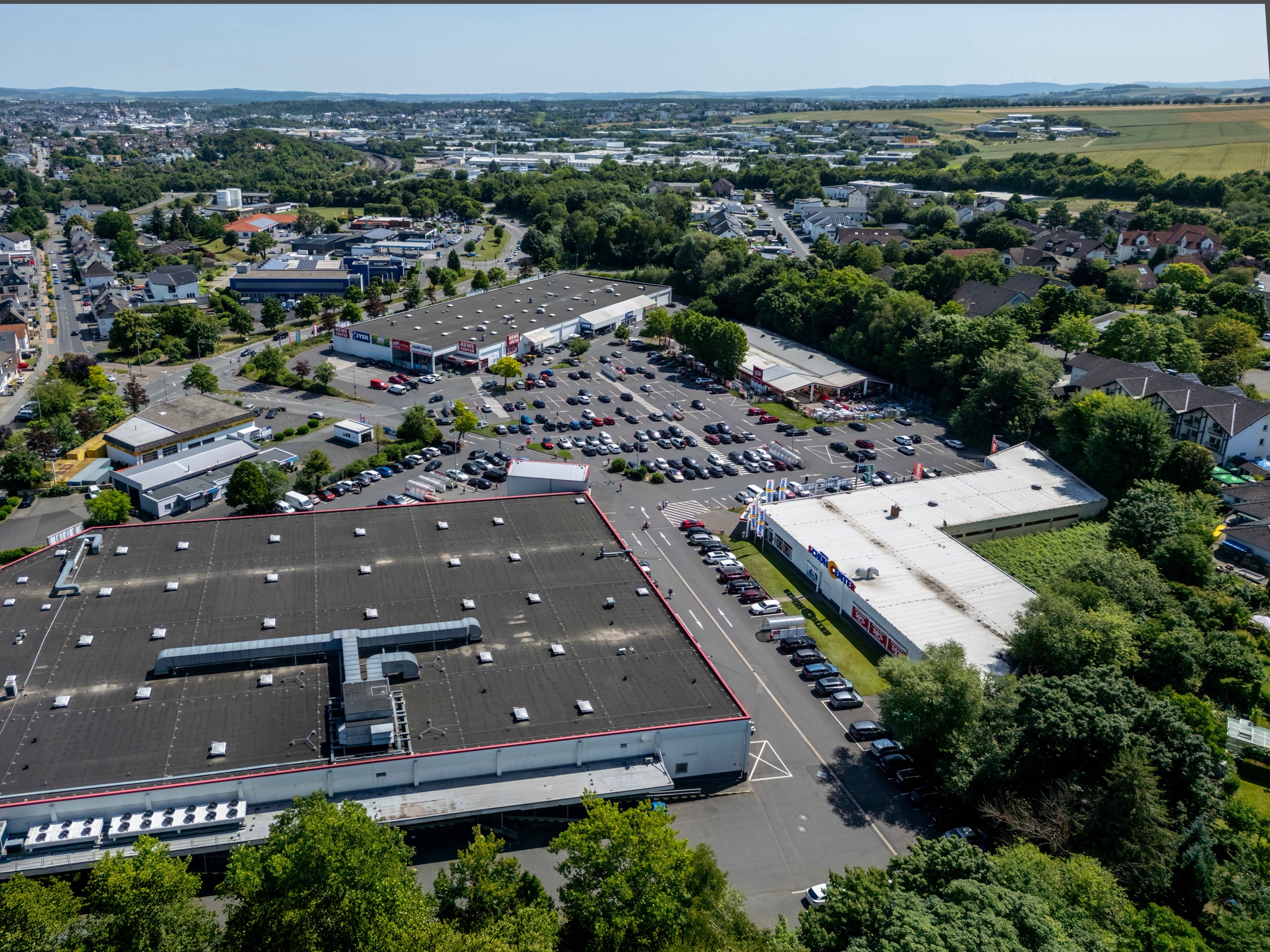 Luftaufnahme eines großen Einkaufszentrums mit mehreren Gebäuden, einem belebten Parkplatz voller Autos und umliegenden Grünflächen. Im Hintergrund sind Wohngebiete und offene Felder zu sehen.