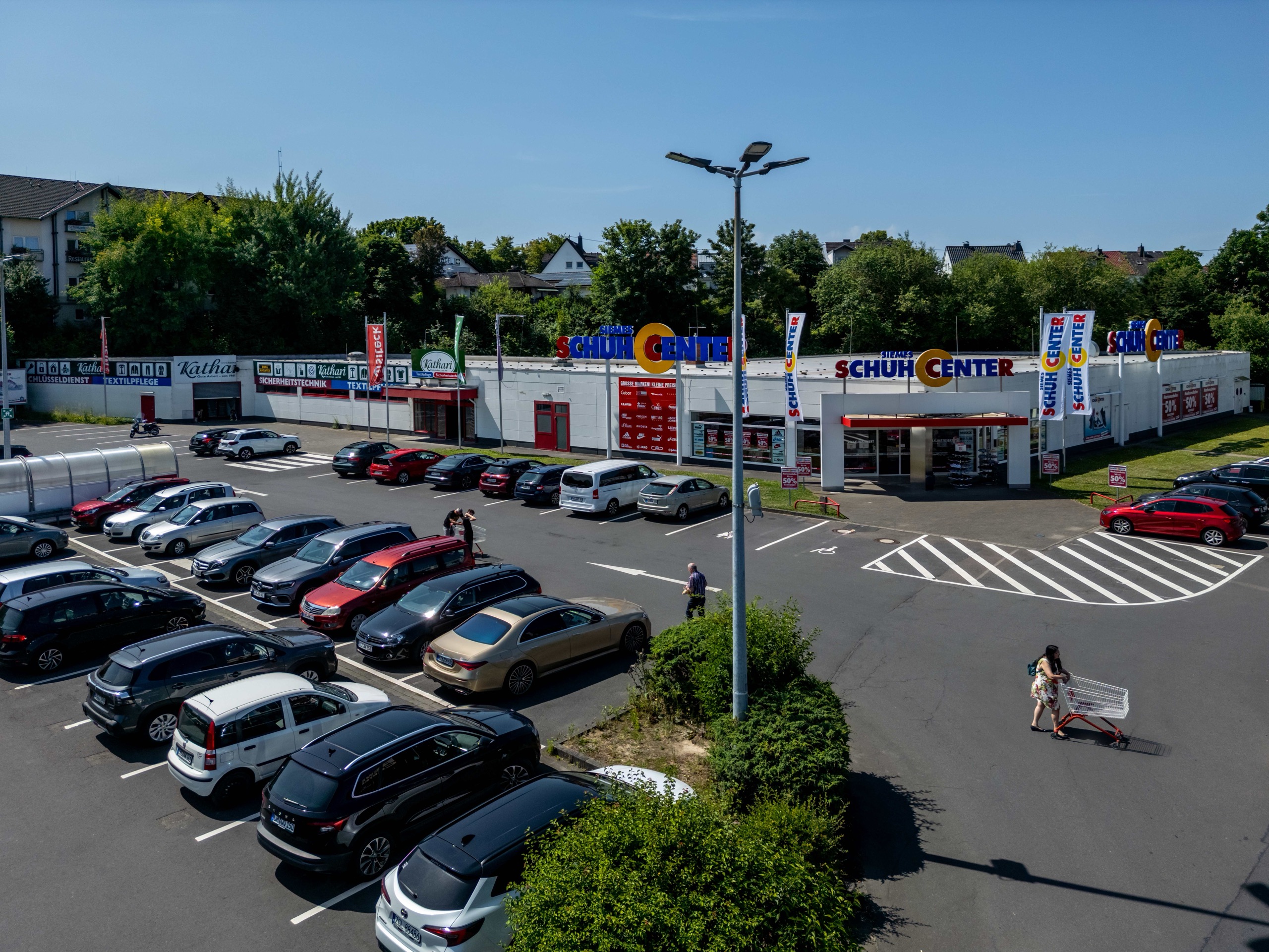 Ein Parkplatz mit mehreren Autos, die vor einem Geschäft namens Schuh Center geparkt sind. Eine Person schiebt einen Einkaufswagen, eine andere geht auf das Geschäft zu. Im Hintergrund sind Bäume und Wohngebäude zu sehen.