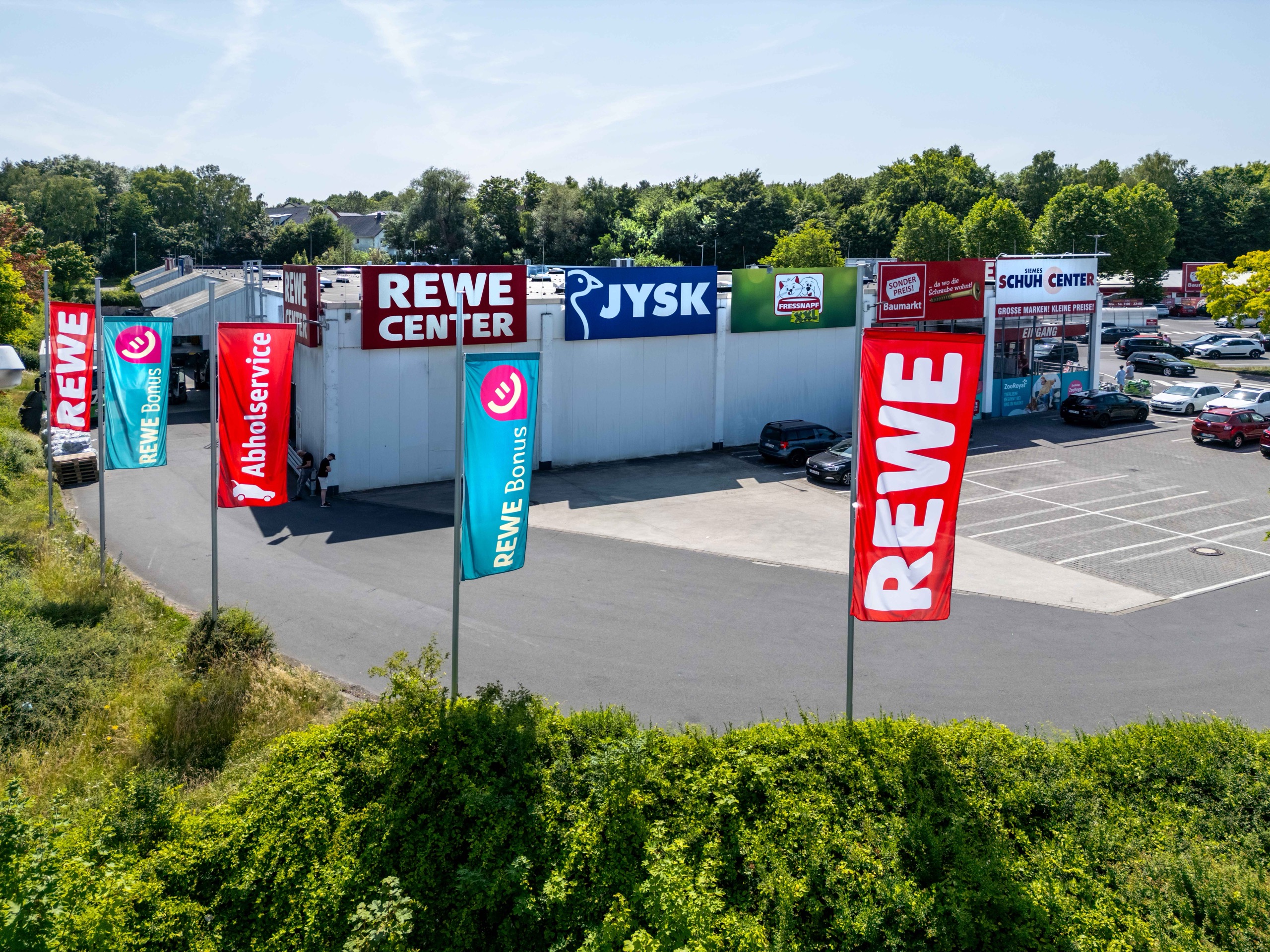 Ein Einkaufszentrum mit Schildern für REWE, JYSK und andere Geschäfte. Rote und blaue REWE-Fahnen stehen auf dem Parkplatz, auf dem mehrere Autos geparkt sind. Bäume und Grünflächen umgeben das Gebiet unter einem teilweise bewölkten Himmel.