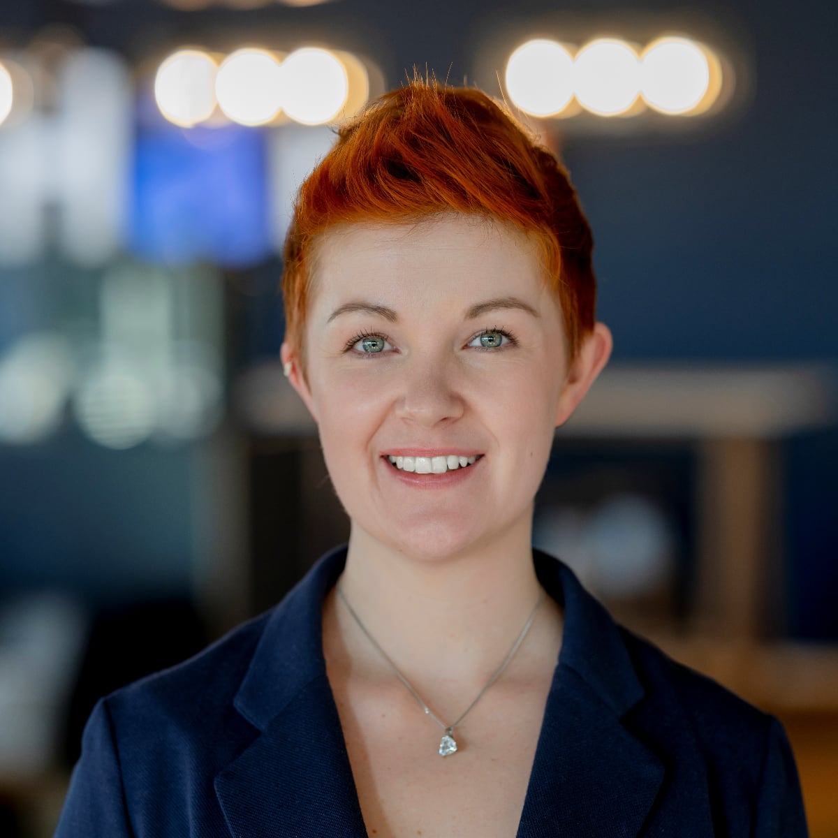 Eine Person mit kurzen roten Haaren, die einen dunklen Blazer und eine Halskette trägt, lächelt, während sie in einem Innenraum mit verschwommenen Lichtern und einem blauen Hintergrund im Hintergrund steht.