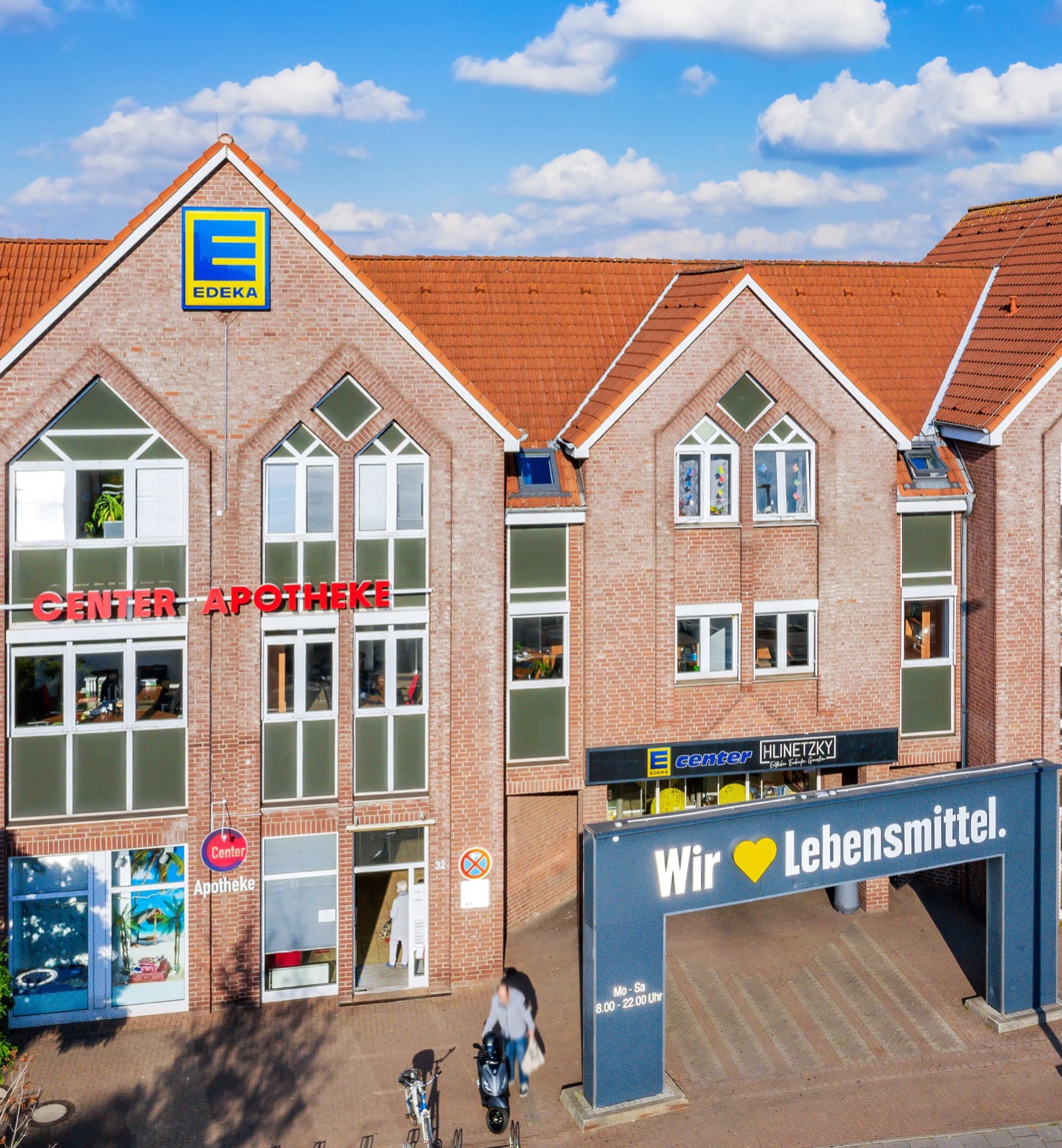Ein Backsteingebäude mit einem EDEKA-Supermarkt und einer Apotheke. Das EDEKA-Schild befindet sich über dem Eingang, und auf einem großen Banner steht Wir ♥ Lebensmittel. Zwei Personen mit Fahrrädern stehen in der Nähe des Eingangs. Der Himmel ist blau mit Wolken.