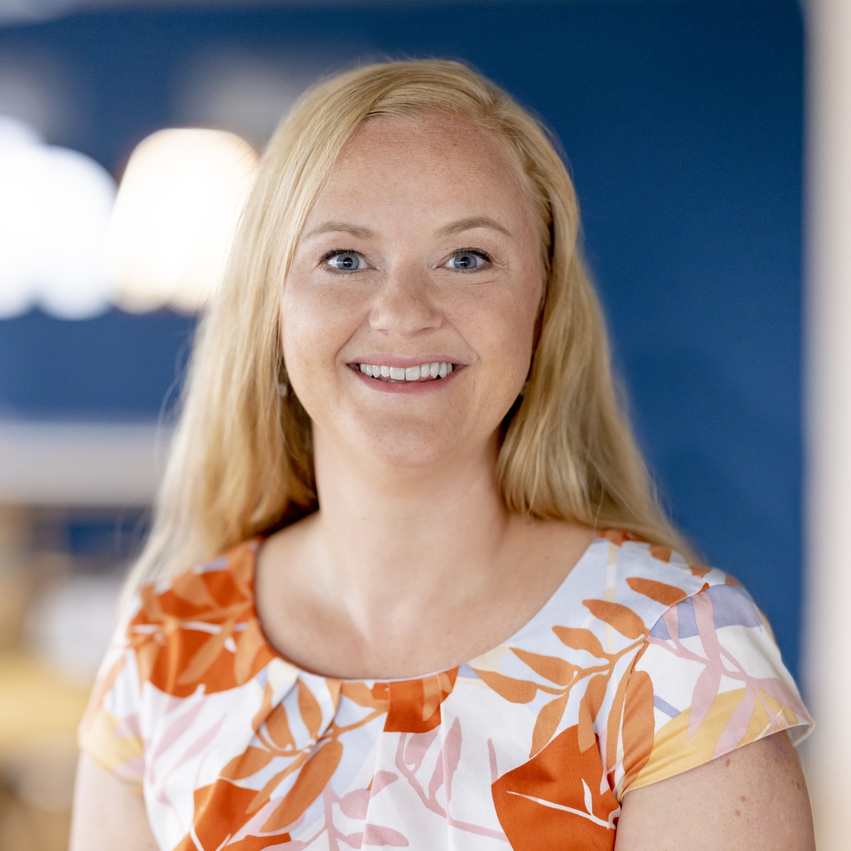 Eine lächelnde Frau mit langen blonden Haaren trägt ein weißes Kleid mit einem orangefarbenen und rosafarbenen Blattmuster und steht in einem Innenraum vor einem unscharfen blauen Hintergrund.