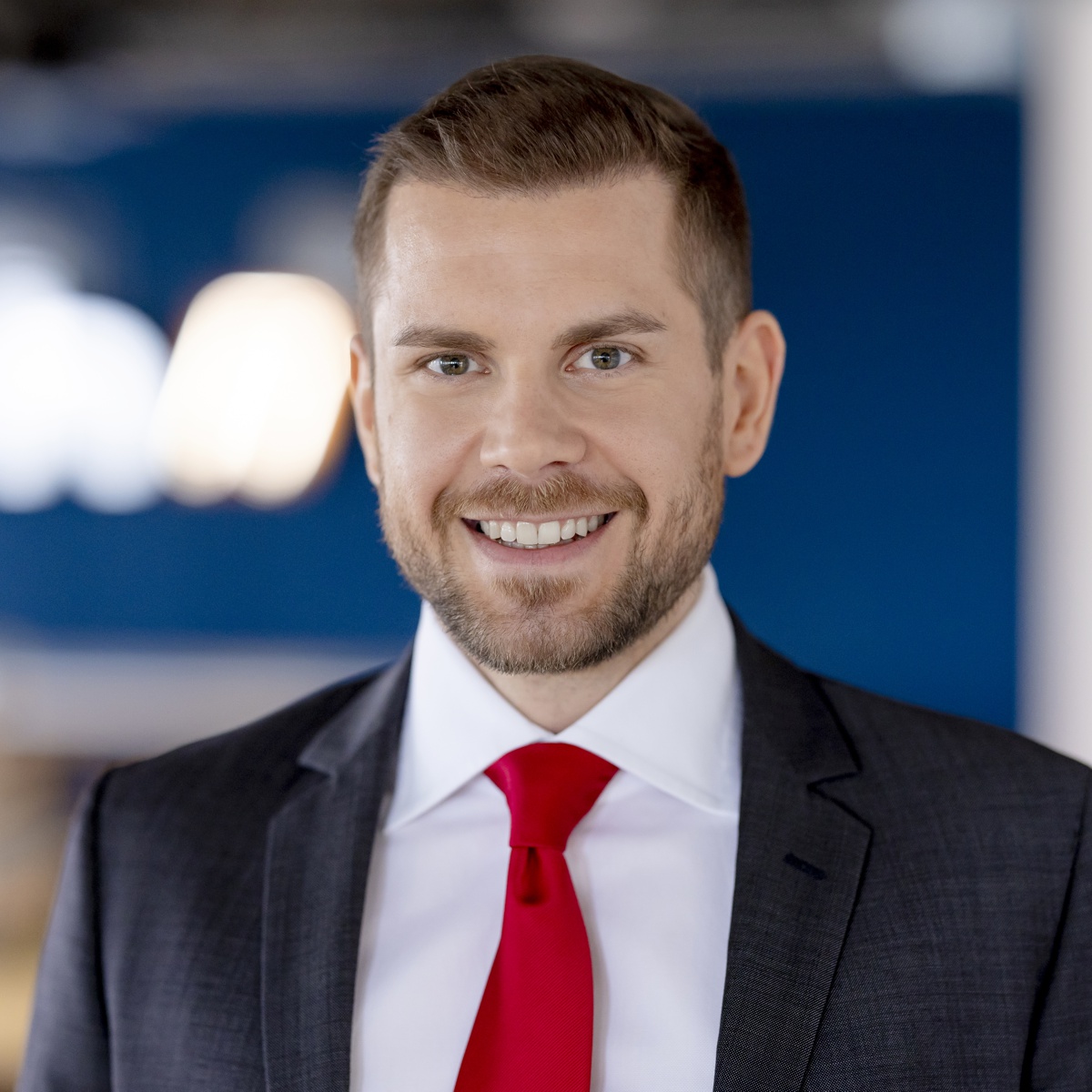 Ein Mann mit kurzen braunen Haaren und einem Bart, der einen dunklen Anzug, ein weißes Hemd und eine rote Krawatte trägt, lächelt in die Kamera vor einem unscharfen blau-weißen Hintergrund.