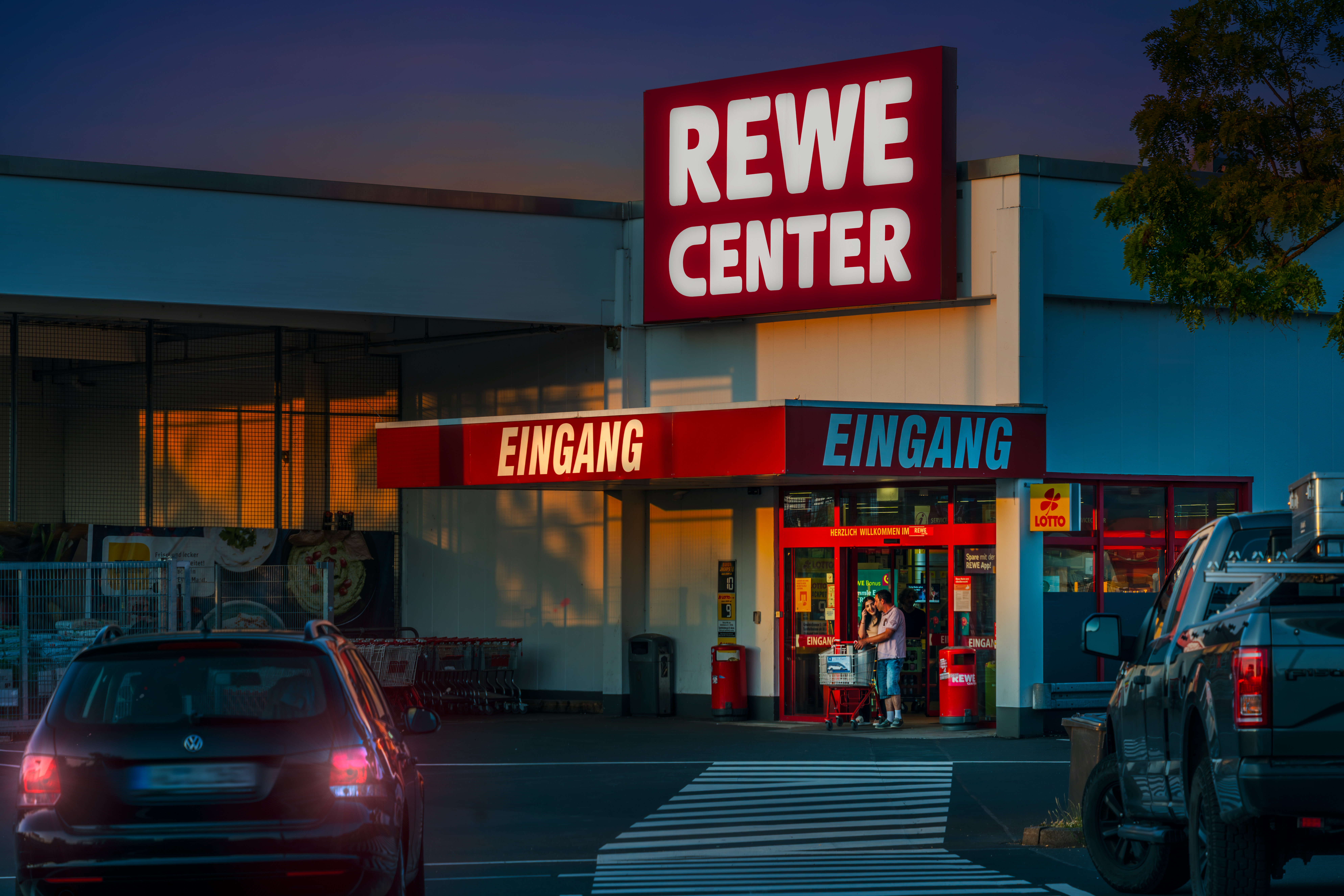 Vor einem REWE-Center-Supermarkt mit roten Eingangsschildern stehen Menschen fahren Autos.
