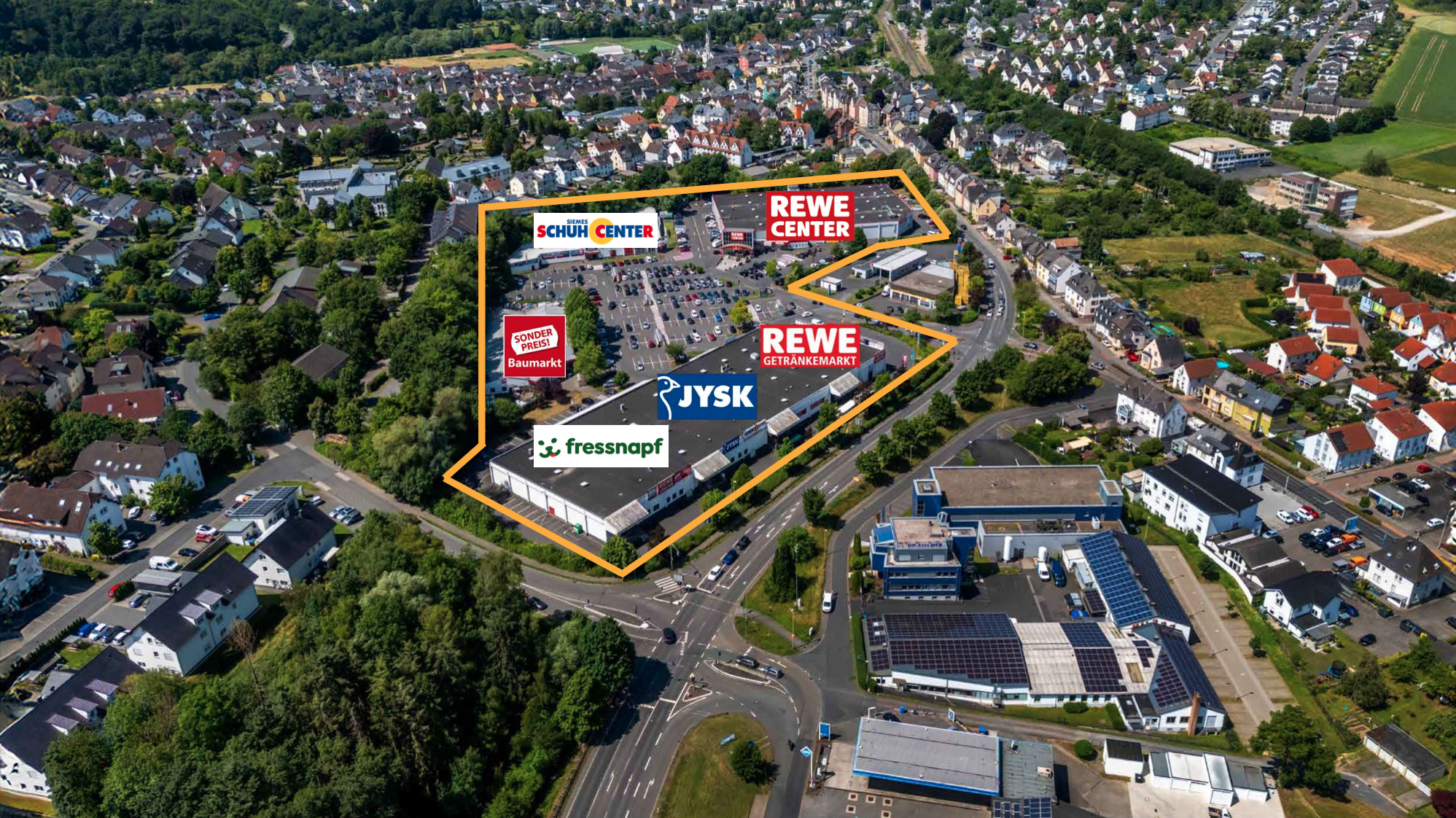 Luftaufnahme eines gelb umrandeten Einkaufszentrums mit Geschäften wie REWE Center, JYSK, Fressnapf und Schuh Center, umgeben von Wohngebieten und Grünflächen in einem Vorstadtviertel.