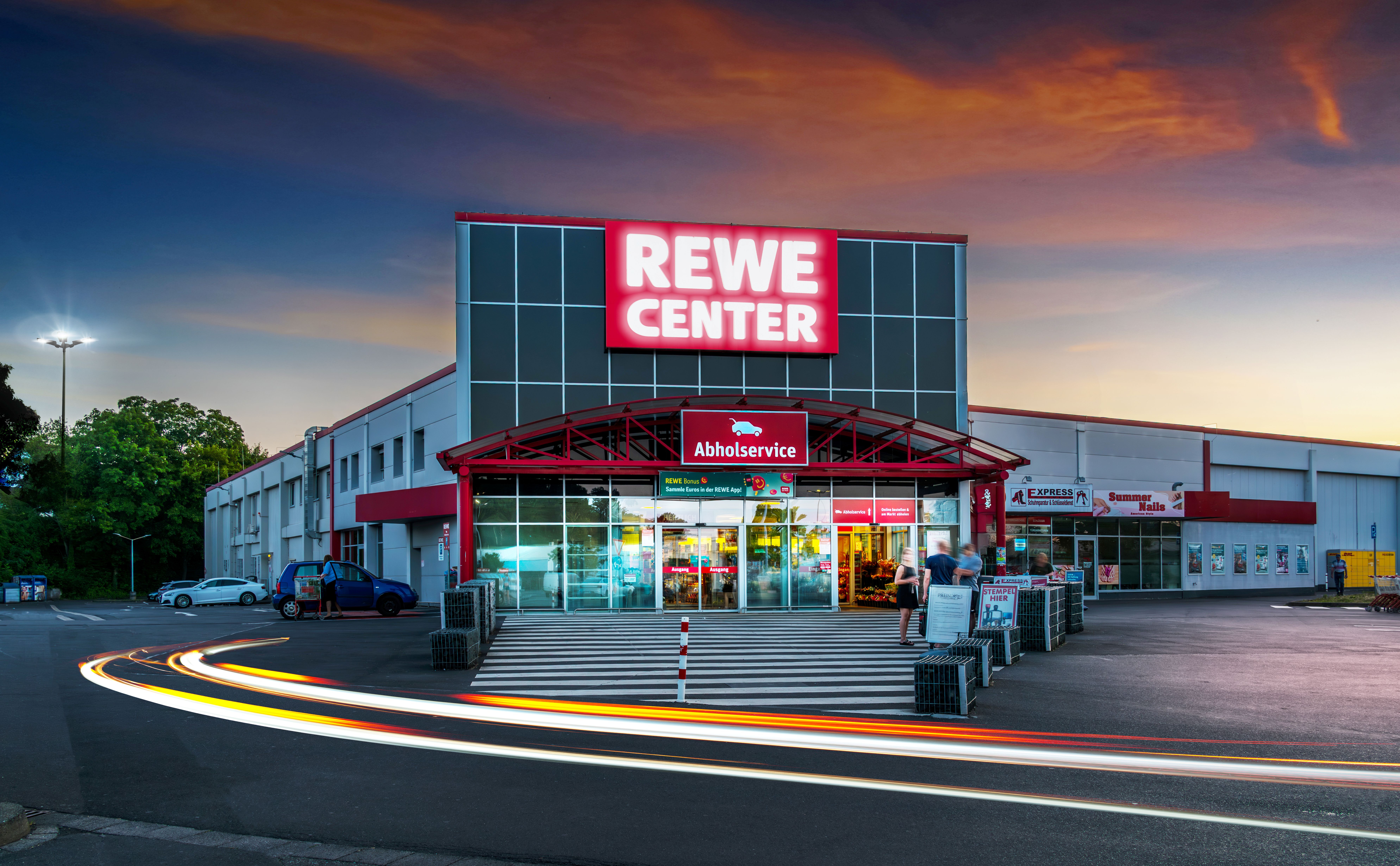 Ein REWE-Center-Supermarkt in der Abenddämmerung mit beleuchteter Beschilderung, ein paar geparkten Autos und Lichtspuren von vorbeifahrenden Fahrzeugen vor dem Eingang.