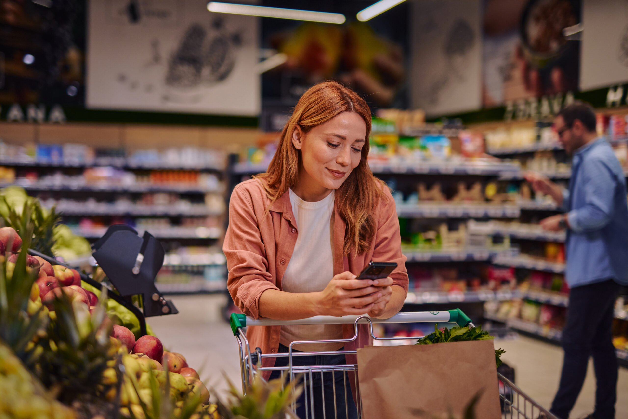 Eine rothaarige Frau steht hinter einem Einkaufswagen in einem Lebensmittelgeschäft, schaut auf ihr Telefon und lächelt. Der Wagen enthält eine Papiertüte und frisches Obst und Gemüse; im Hintergrund sind Regale mit Lebensmitteln zu sehen.