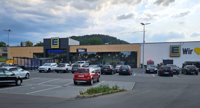 Ein Supermarktgebäude mit EDEKA-Schildern, parkende Autos davor, ein teilweise bewölkter Himmel darüber und ein kleiner Hügel im Hintergrund. In der Nähe des Eingangs sind Einkäufer zu sehen.