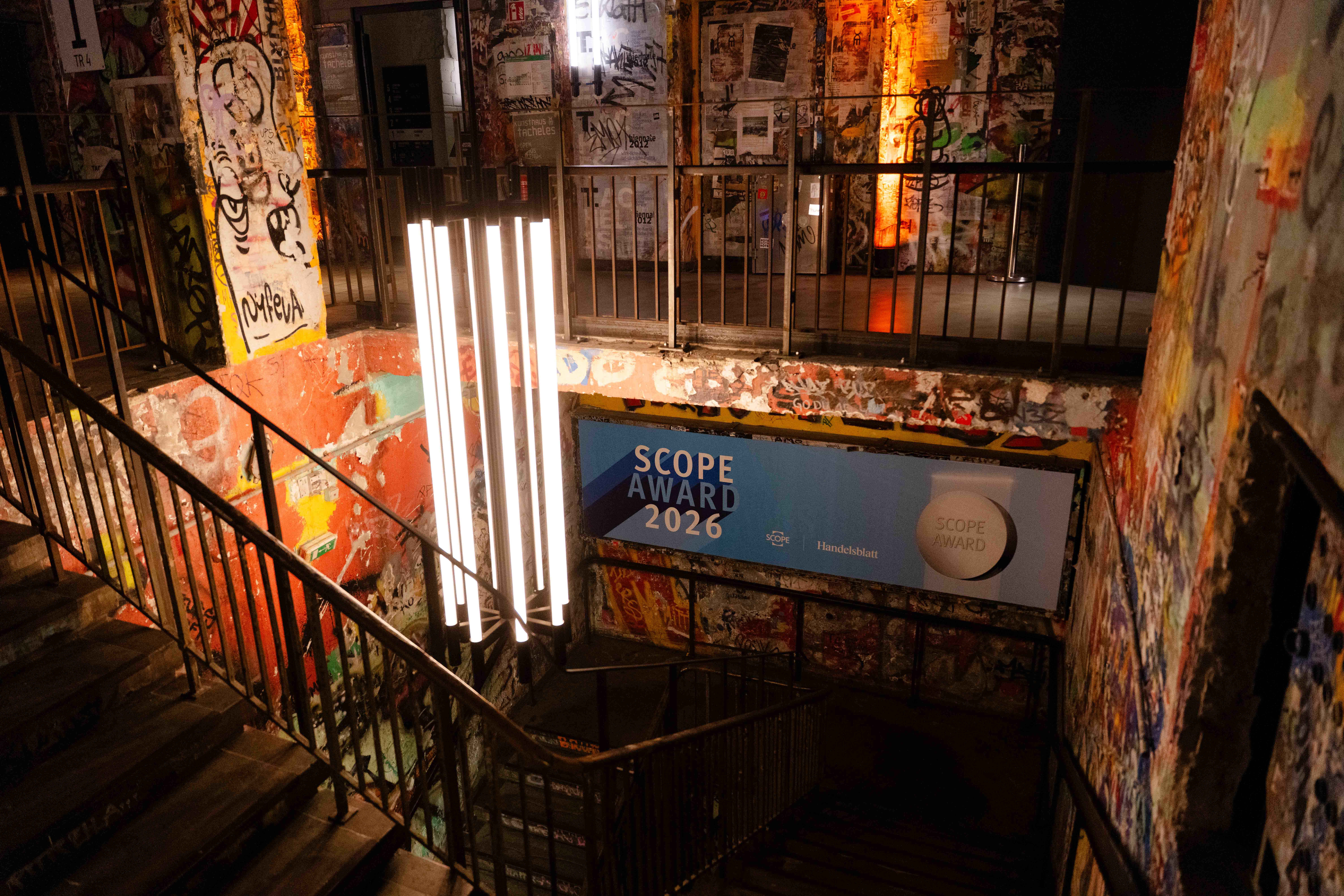 Ein schwach beleuchtetes, mit Graffiti bedecktes Treppenhaus mit einem vertikalen Kronleuchter und einem großen blauen Schild mit der Aufschrift SCOPE AWARD 2026 an der Wand. Mehrere Treppen und Geländer sind auf dem Bild zu sehen.