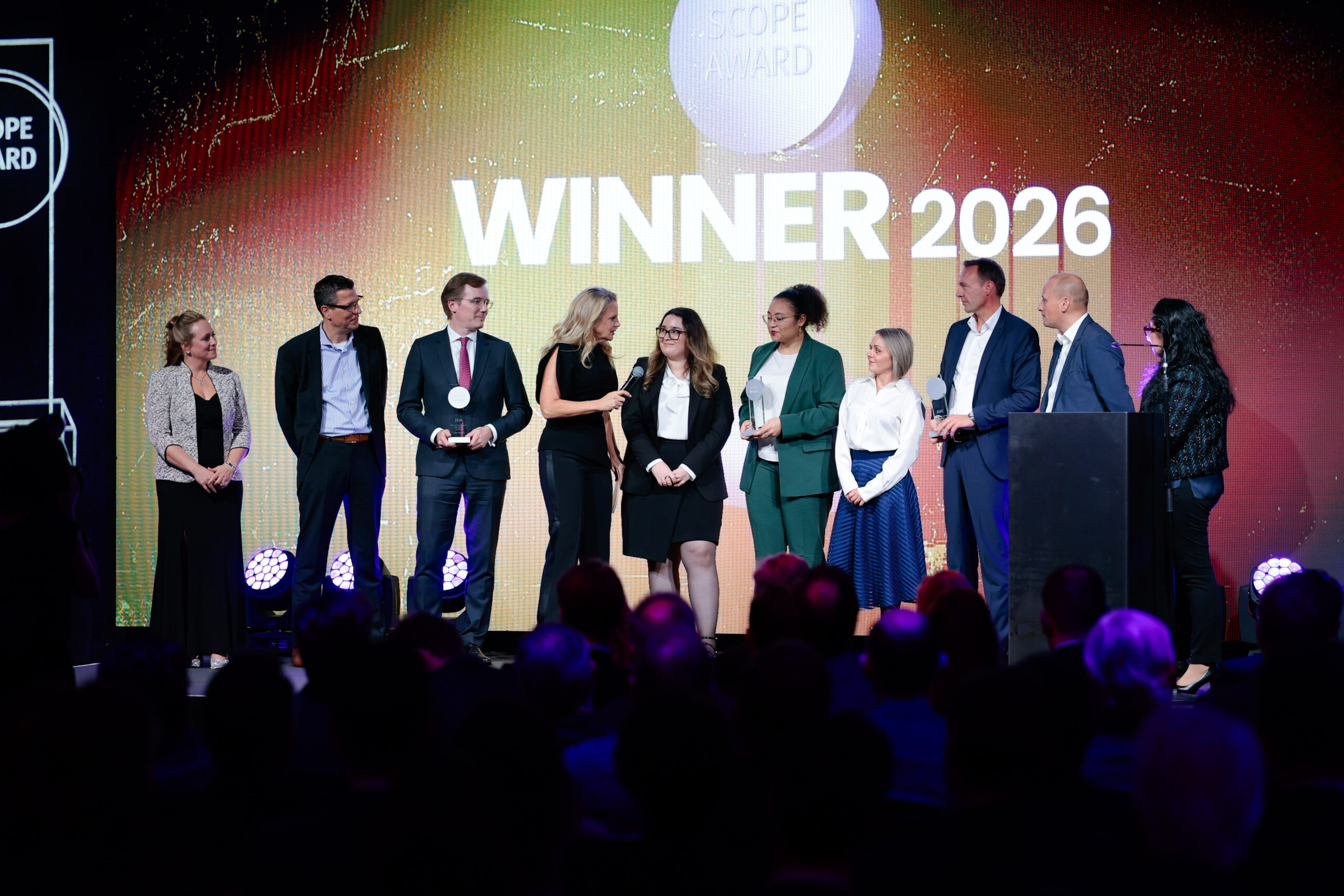 Eine Gruppe von zehn Personen steht unter hellem Licht auf der Bühne und blickt auf das Publikum. Hinter ihnen steht in großen Lettern "WINNER 2026" und "SCOPE AWARD". Einige halten Mikrofone, und alle scheinen eine Leistung zu feiern.