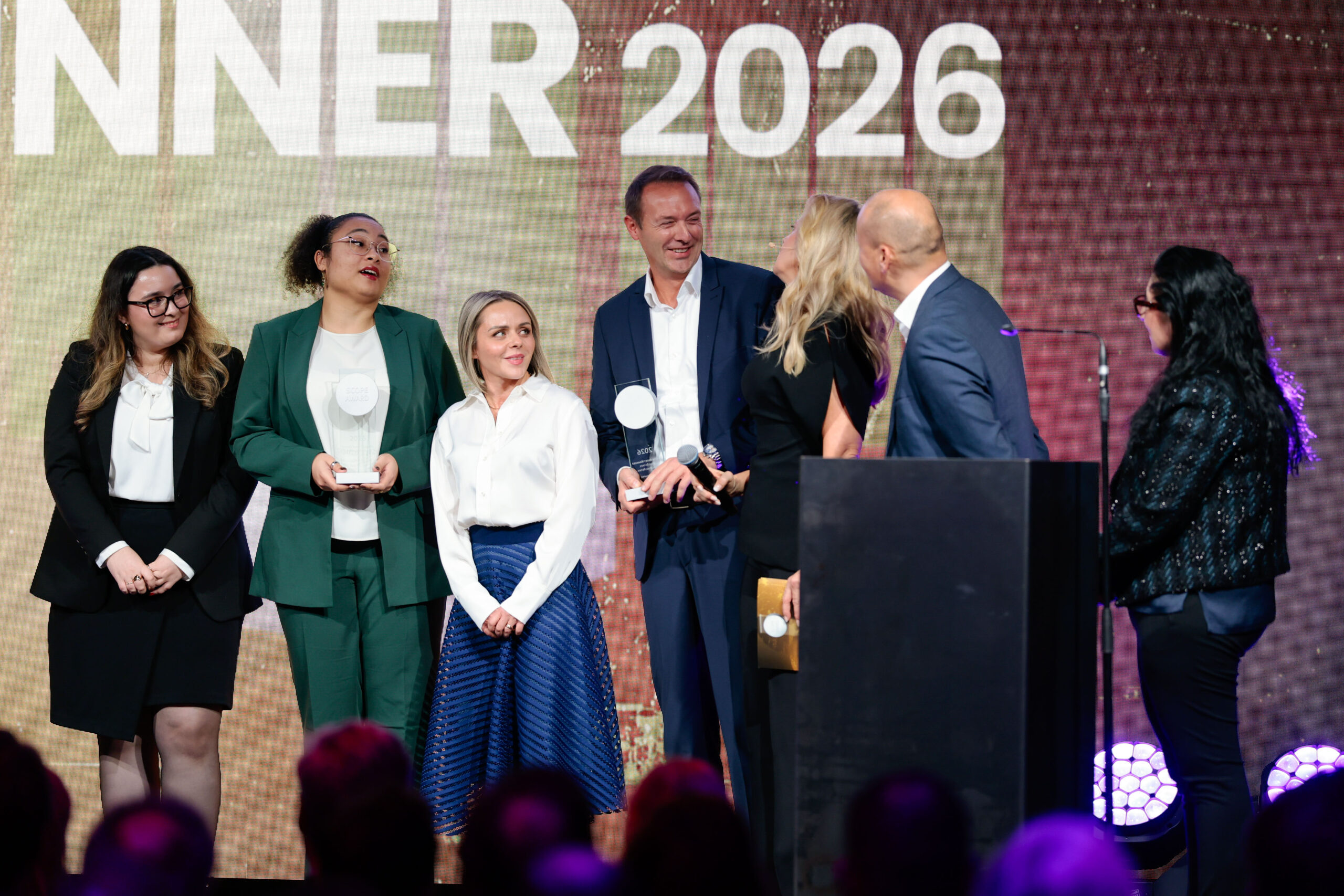 Eine Gruppe von sechs Personen steht bei einer Preisverleihung auf der Bühne, drei von ihnen halten Trophäen. Im Hintergrund ist WINNER 2026 zu sehen. Zwei Personen auf dem Podium scheinen zu sprechen, während die anderen zusehen und lächeln.