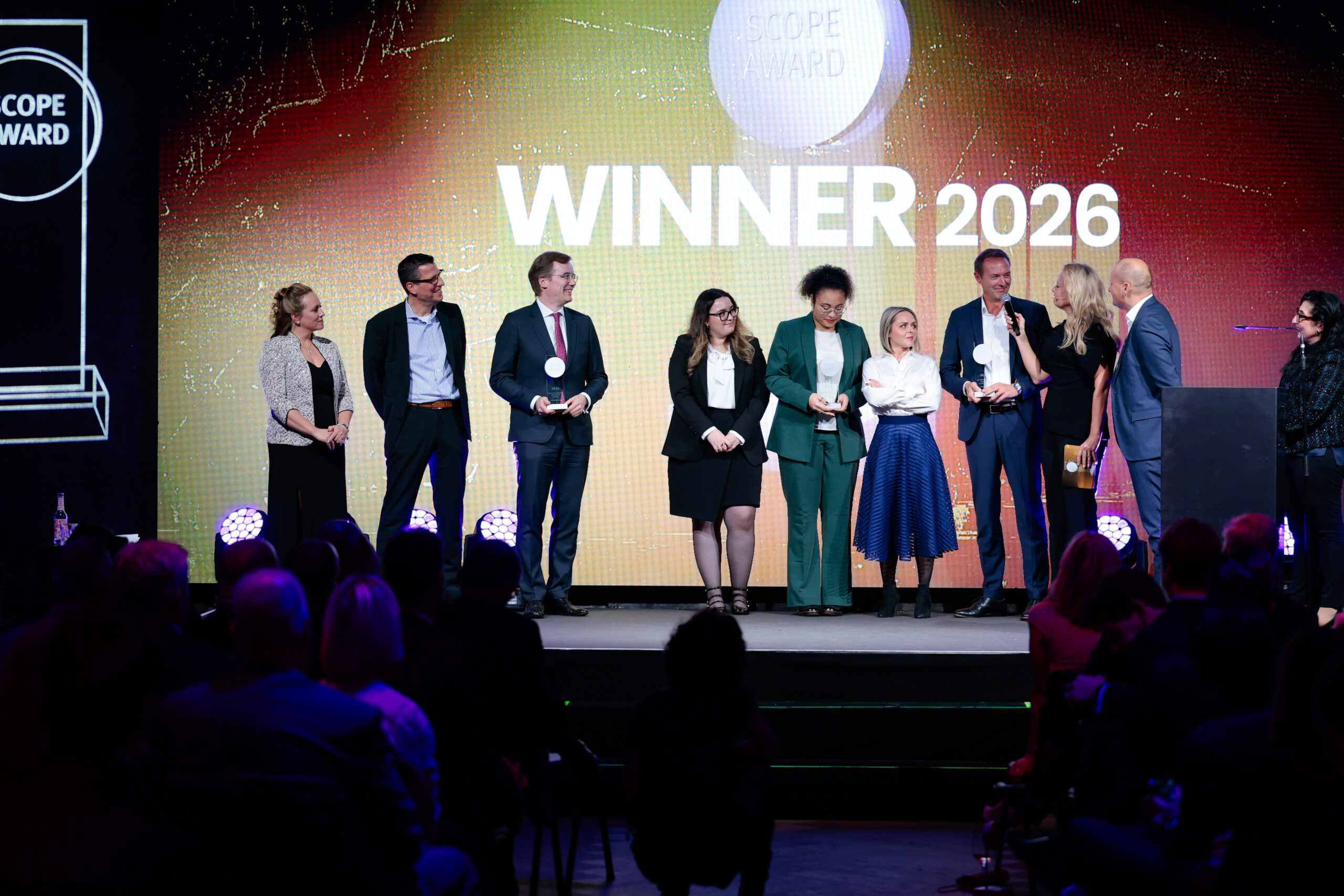 Eine Gruppe von Personen steht auf der Bühne und nimmt bei einer Veranstaltung Auszeichnungen entgegen. Im Hintergrund sind WINNER 2026 und eine Preisgrafik zu sehen. Im Vordergrund sitzt ein Publikum, das die Zeremonie verfolgt.