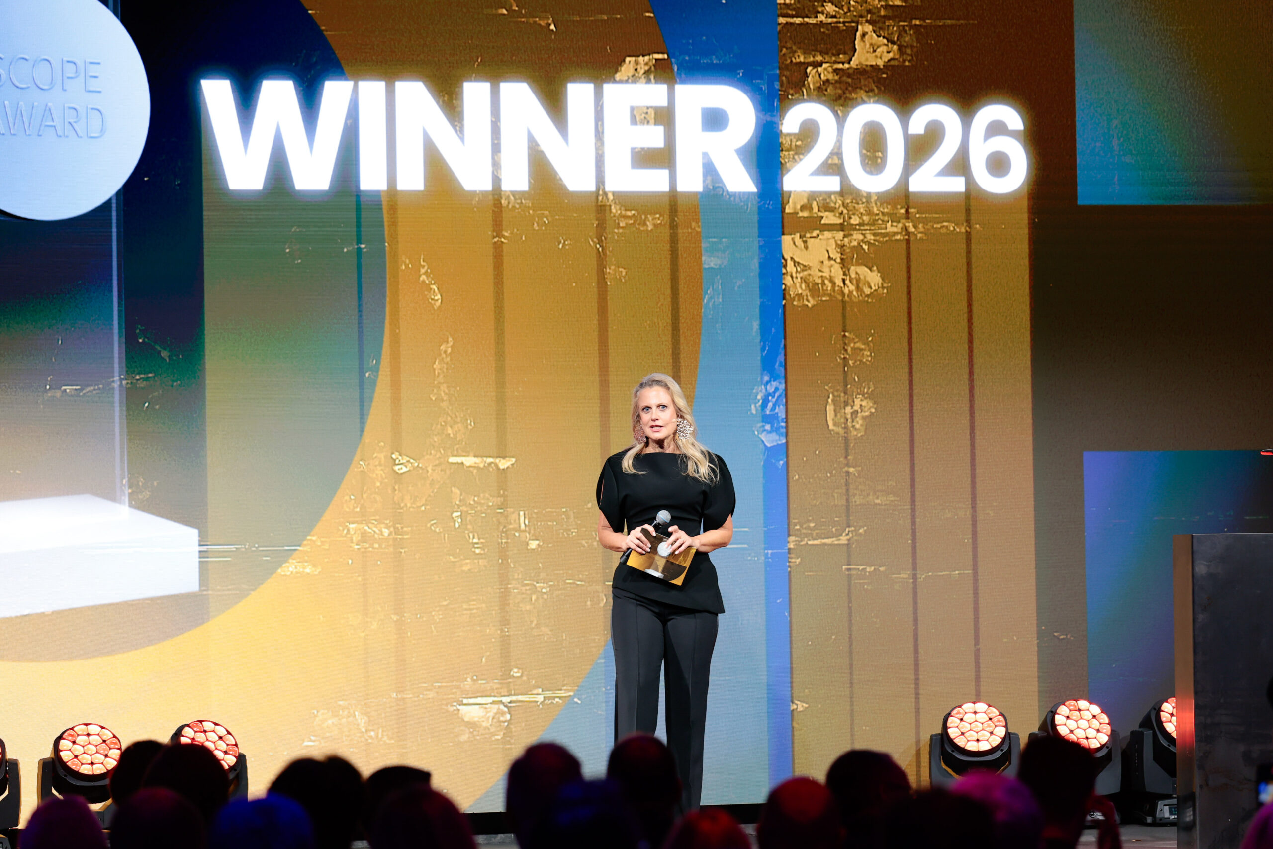 Eine Frau steht auf der Bühne, hält eine Trophäe in der Hand und spricht, während hinter ihr auf einem großen Bildschirm in fetten weißen Buchstaben "WINNER 2026" zu lesen ist.