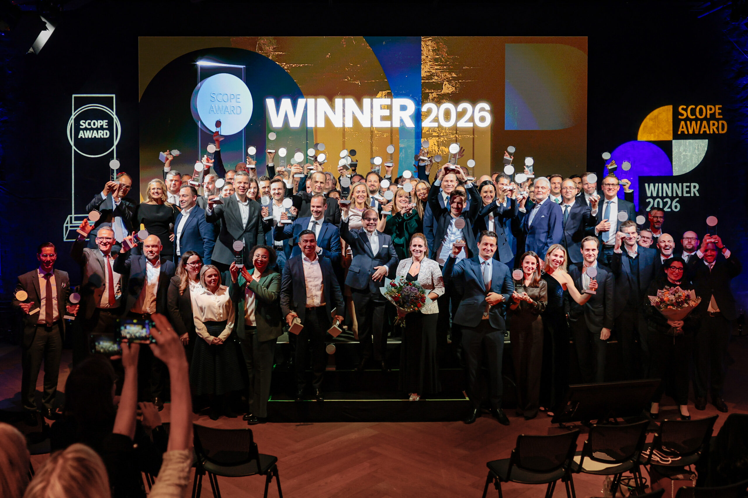 Eine große Gruppe formell gekleideter Menschen steht auf der Bühne, hält Trophäen und Blumen in der Hand und feiert den Scope Award Winner 2026 unter hellen Lichtern mit bunten Preisschildern im Hintergrund.
