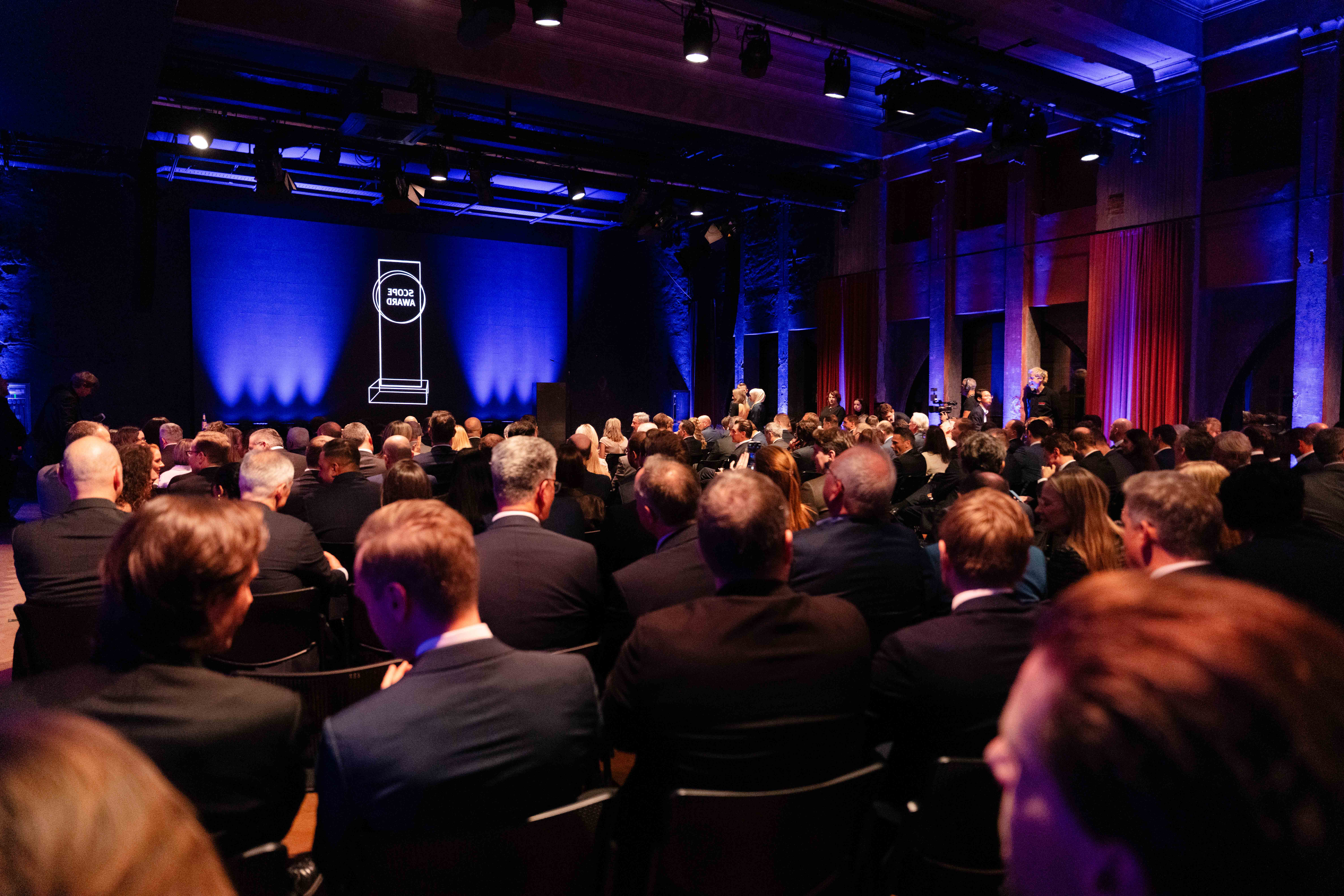 Ein großes Publikum in förmlicher Kleidung sitzt vor einer Bühne mit einem beleuchteten Bildschirm, auf dem eine Preisgrafik in einem schwach beleuchteten Veranstaltungsraum angezeigt wird.