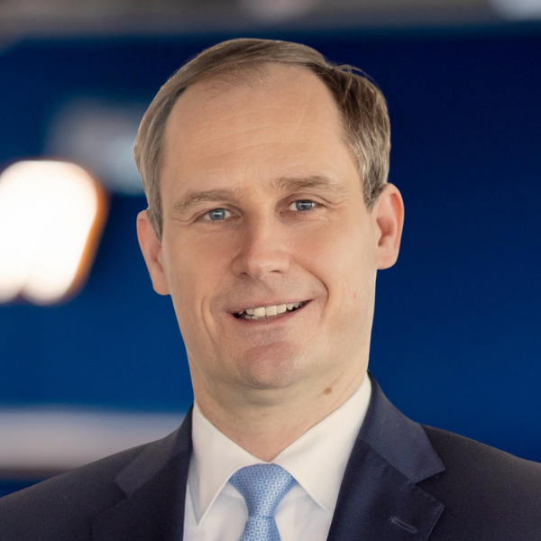 Ein Mann im Anzug und mit hellblauer Krawatte lächelt in die Kamera vor einem unscharfen blauen Hintergrund.