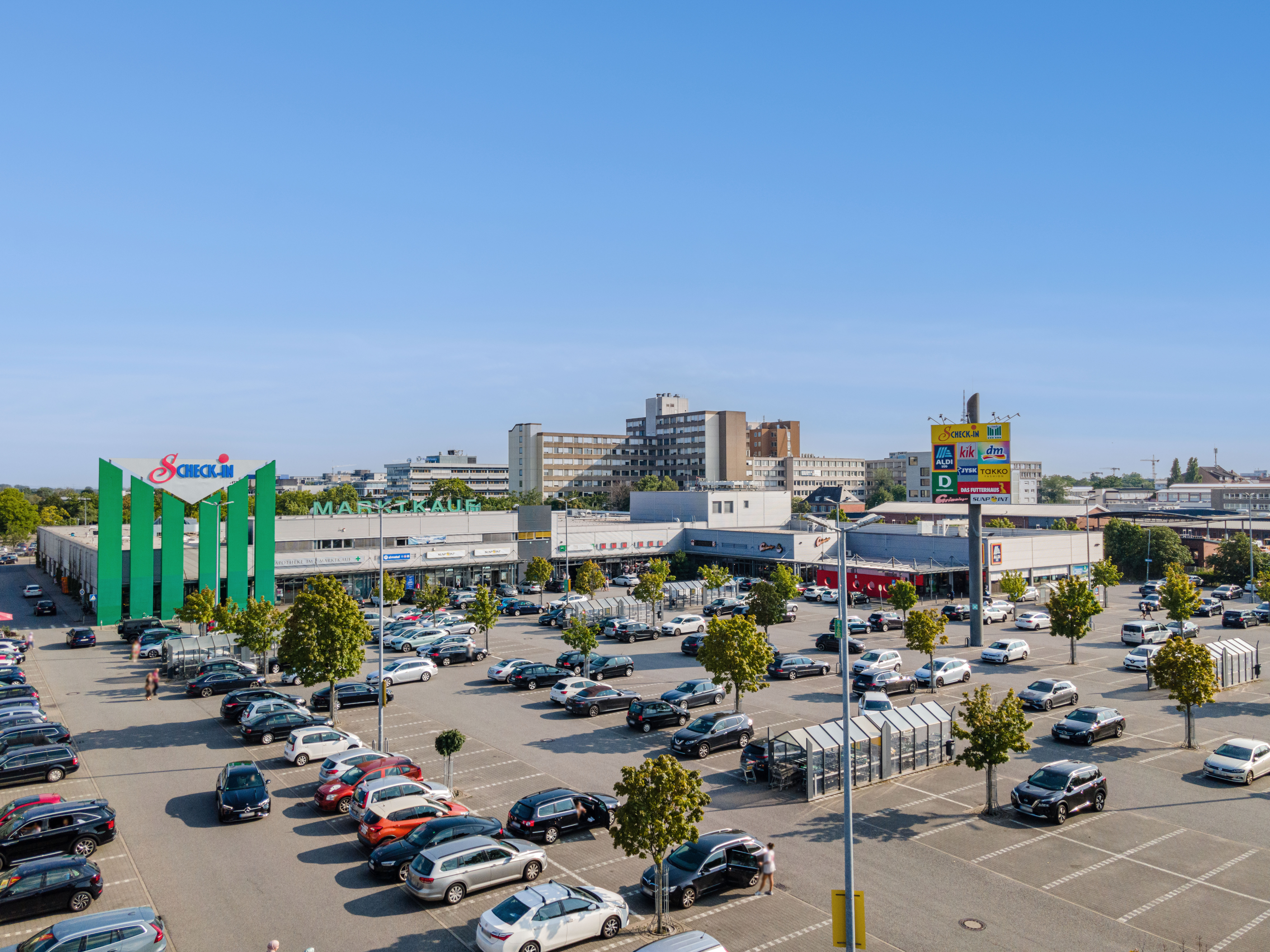 Ein großer Parkplatz mit vielen Autos befindet sich vor einem Einkaufszentrum mit grünen vertikalen Akzenten und sichtbaren Ladenschildern unter einem klaren blauen Himmel. Im Hintergrund sind mehrere Gebäude zu sehen.