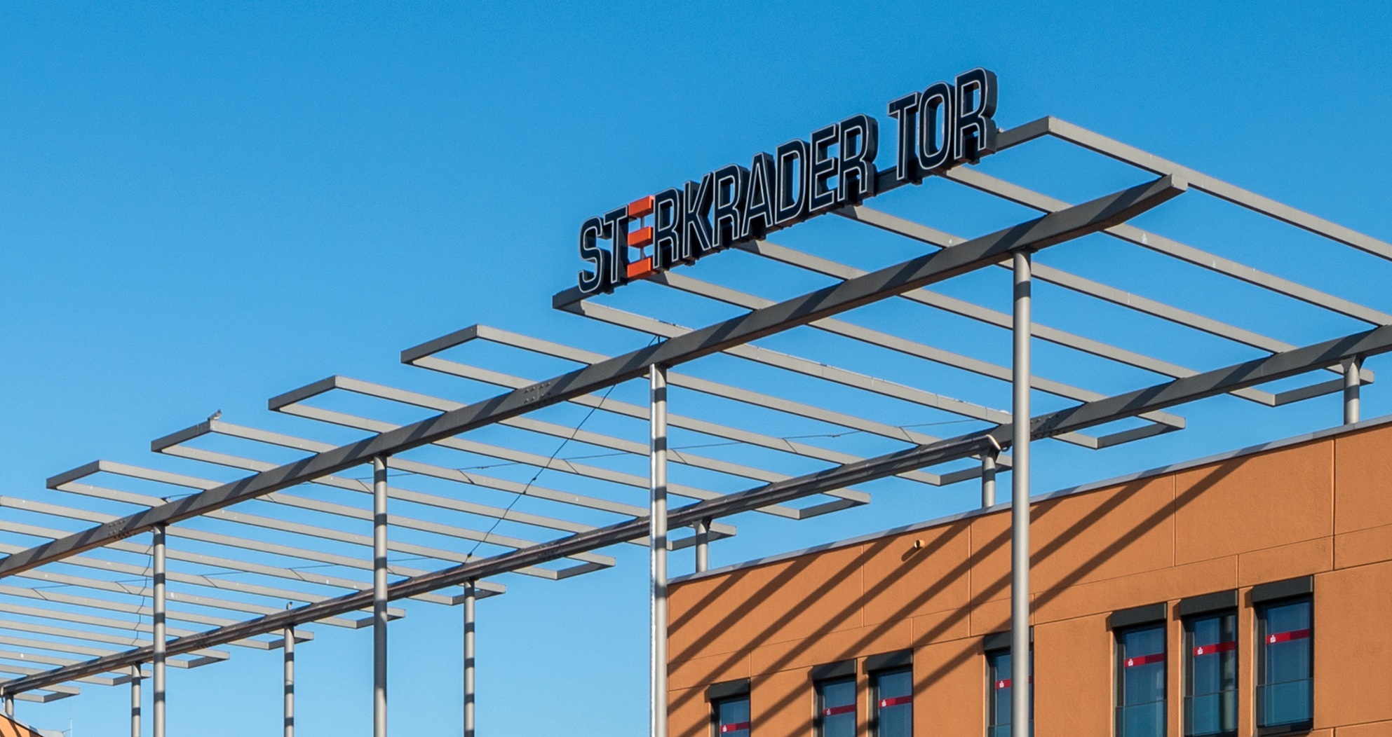 Ein modernes Gebäude mit orangefarbener Fassade, darüber ein Metallgerüst und ein Schild mit der Aufschrift STERRADER TOR vor einem klaren blauen Himmel.