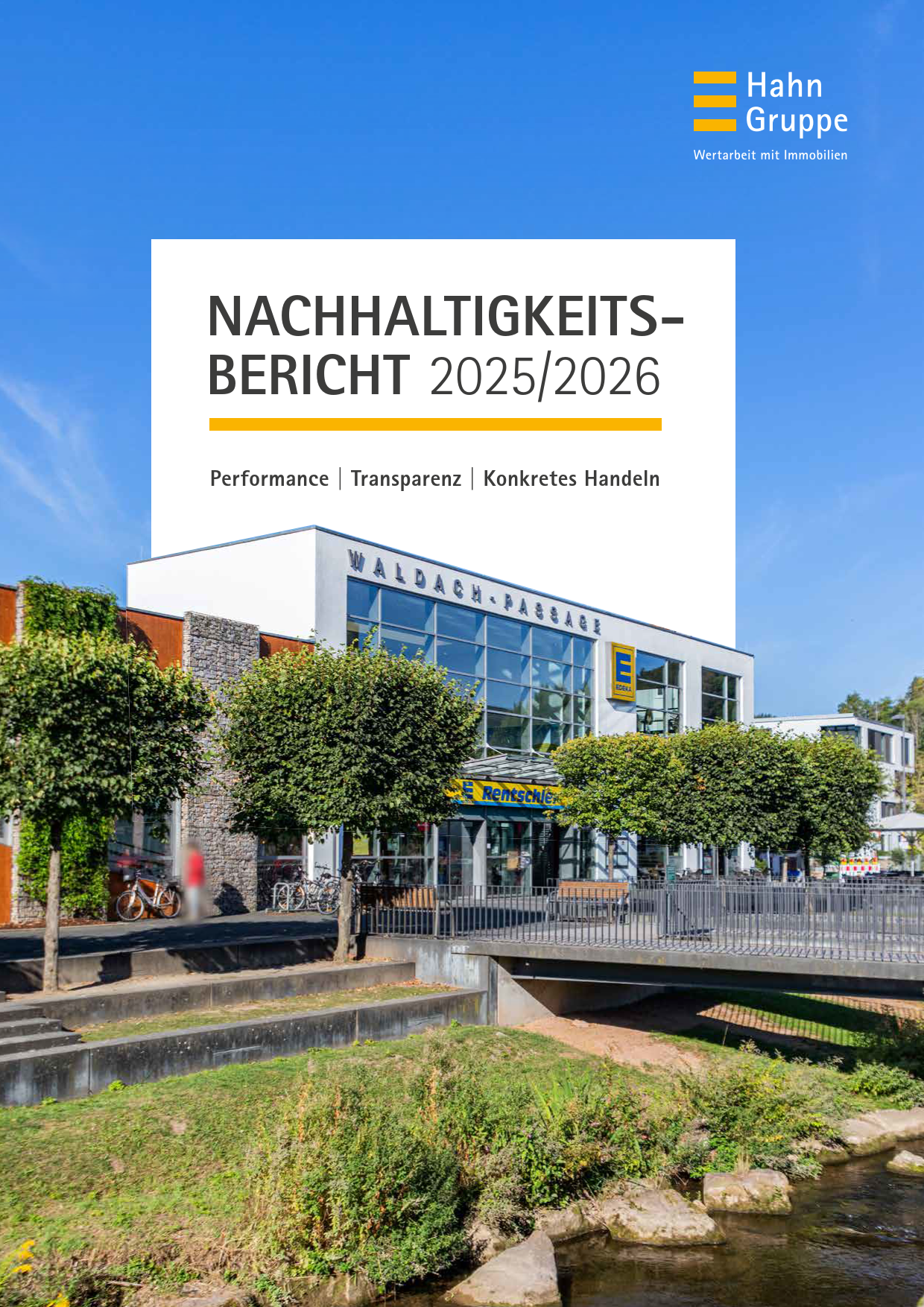 Ein modernes Geschäftsgebäude mit großen Glasfenstern und dem Schriftzug Waldach-Passage, umgeben von Grünflächen, einem kleinen Kanal und einer Fußgängerbrücke, unter einem klaren blauen Himmel. Text: Nachhaltigkeitsbericht 2025/2026.