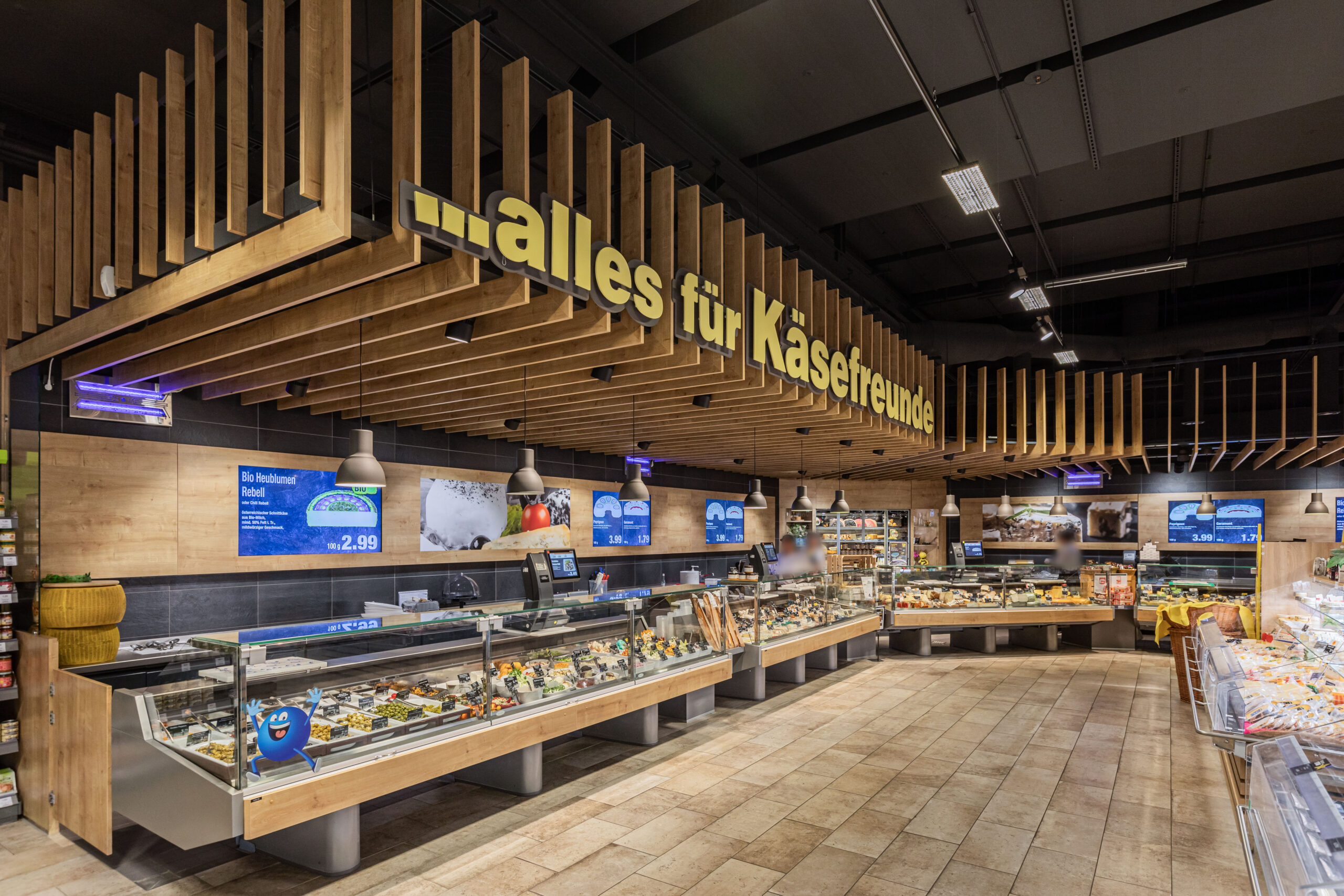 Vor einem REWE-Center-Supermarkt mit roten Eingangsschildern stehen Menschen fahren Autos.