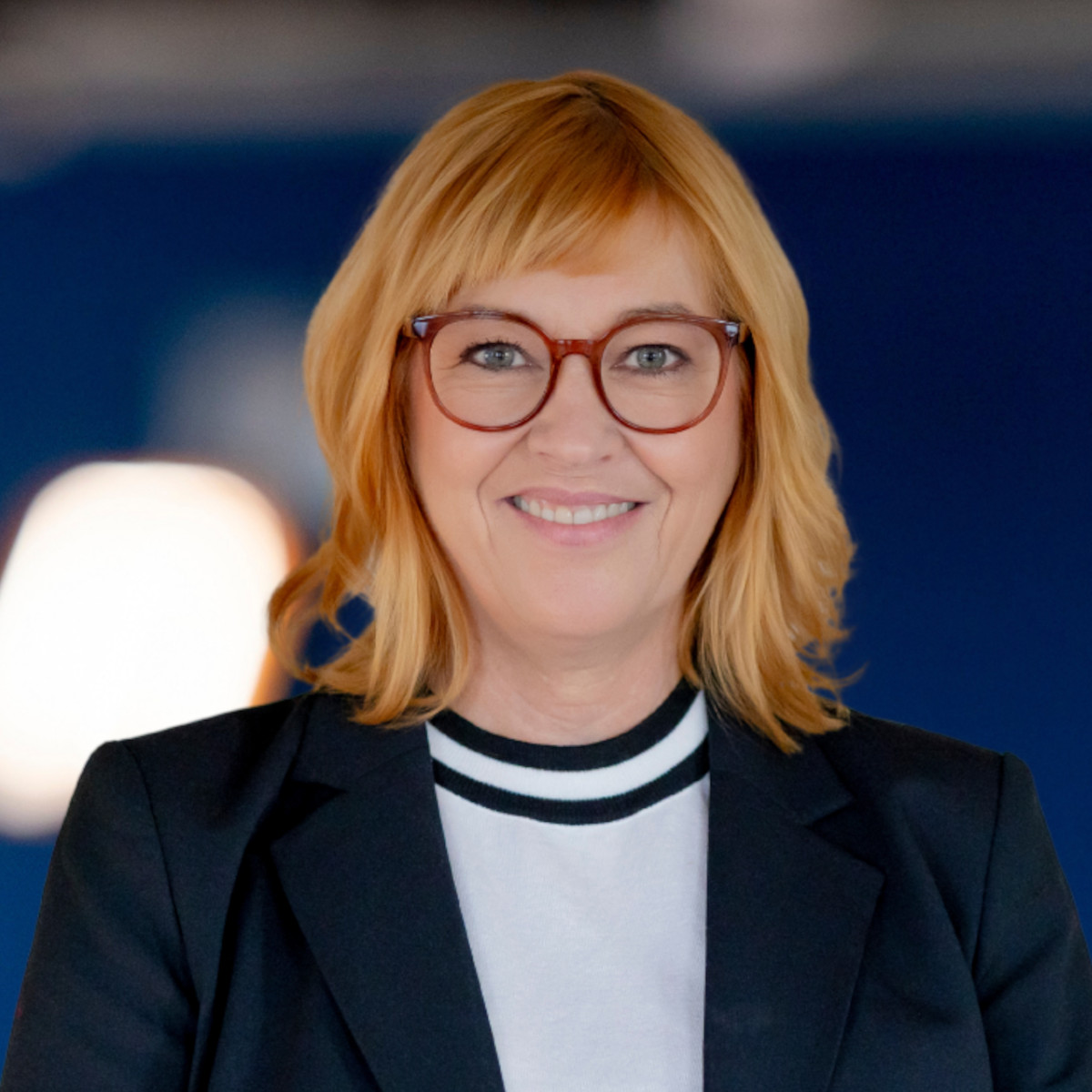 Eine Person mit kurzen grauen Haaren und Brille lächelt in einem dunklen Jackett und einem weißen Hemd. Der Hintergrund ist leicht verschwommen, mit dunkelblauen Tönen und einem Hauch von Licht.