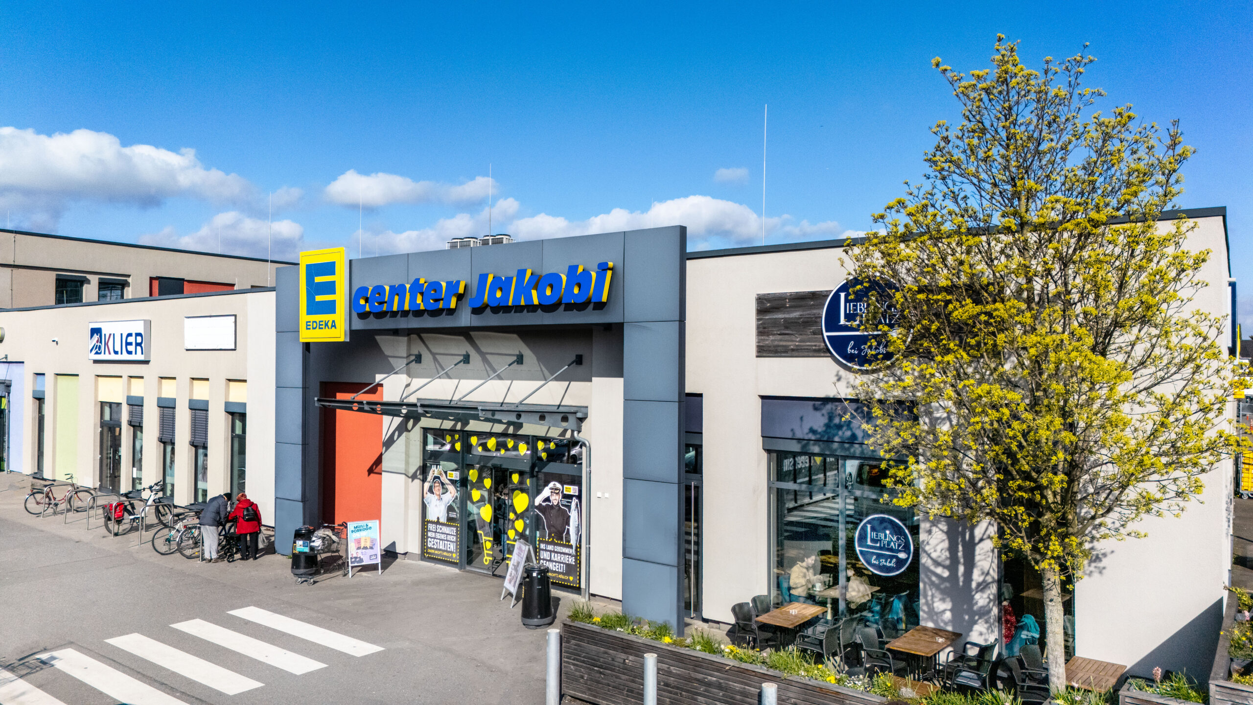Vor einem REWE-Center-Supermarkt mit roten Eingangsschildern stehen Menschen fahren Autos.