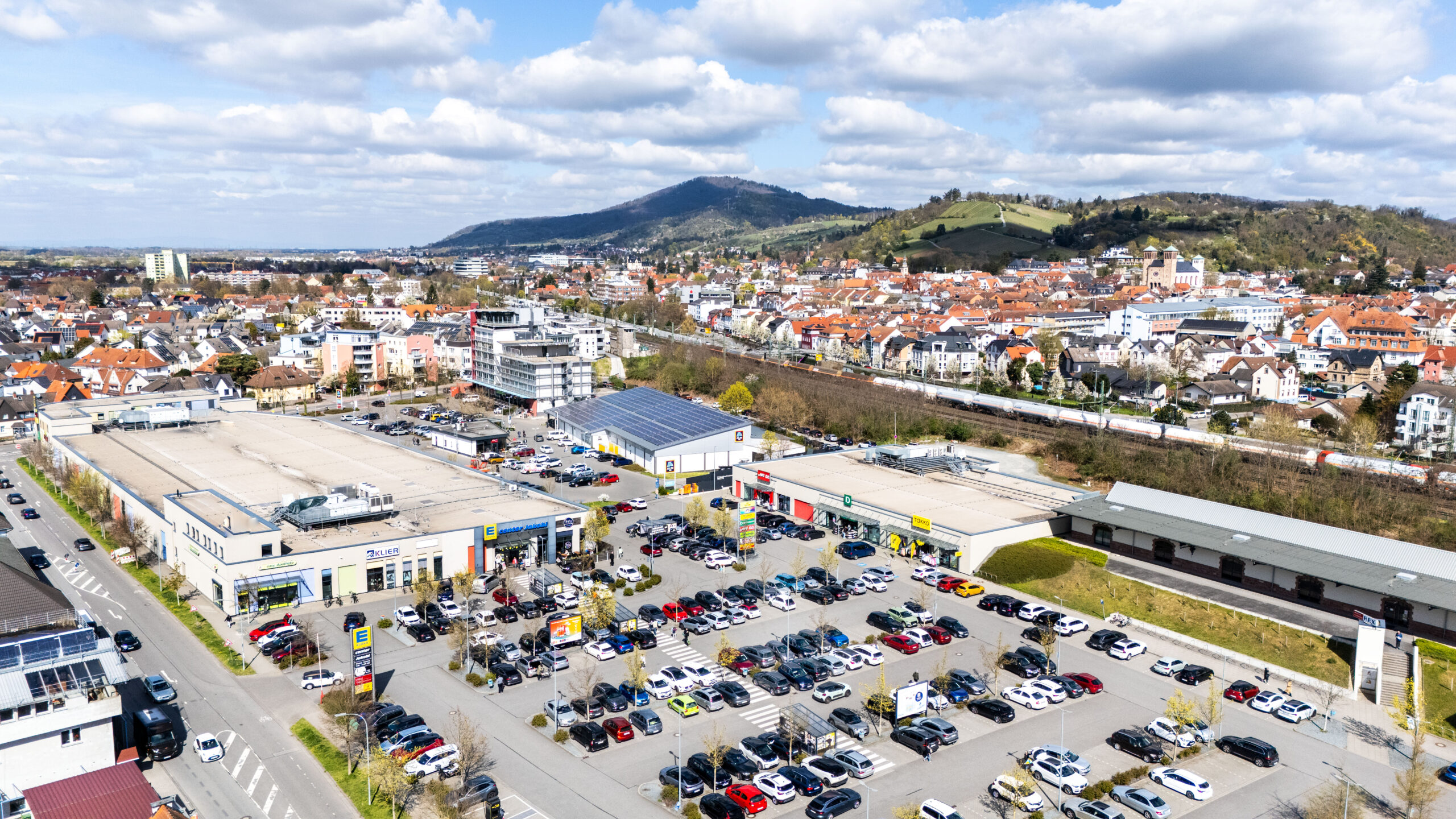 Vor einem REWE-Center-Supermarkt mit roten Eingangsschildern stehen Menschen fahren Autos.