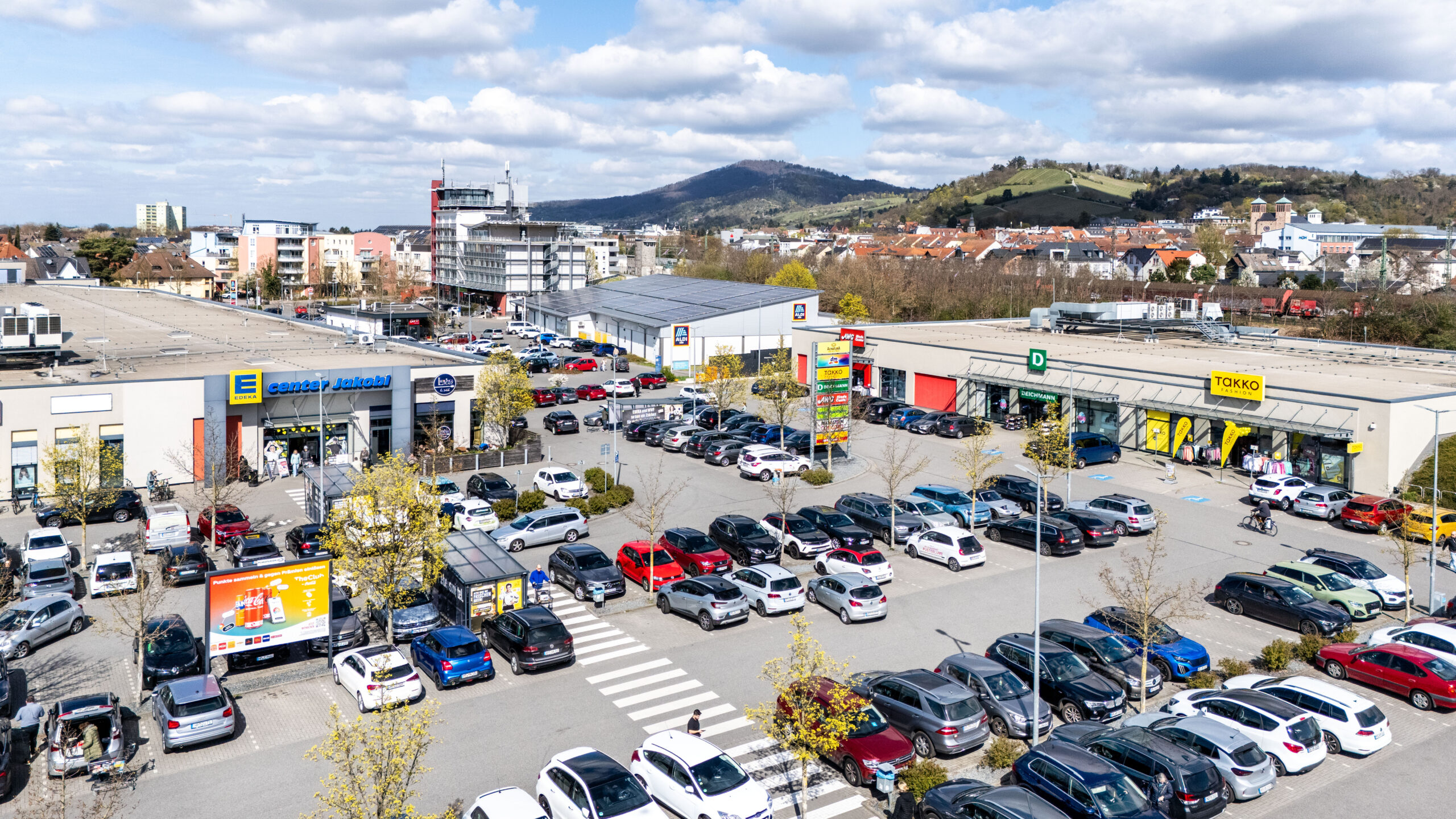 Vor einem REWE-Center-Supermarkt mit roten Eingangsschildern stehen Menschen fahren Autos.
