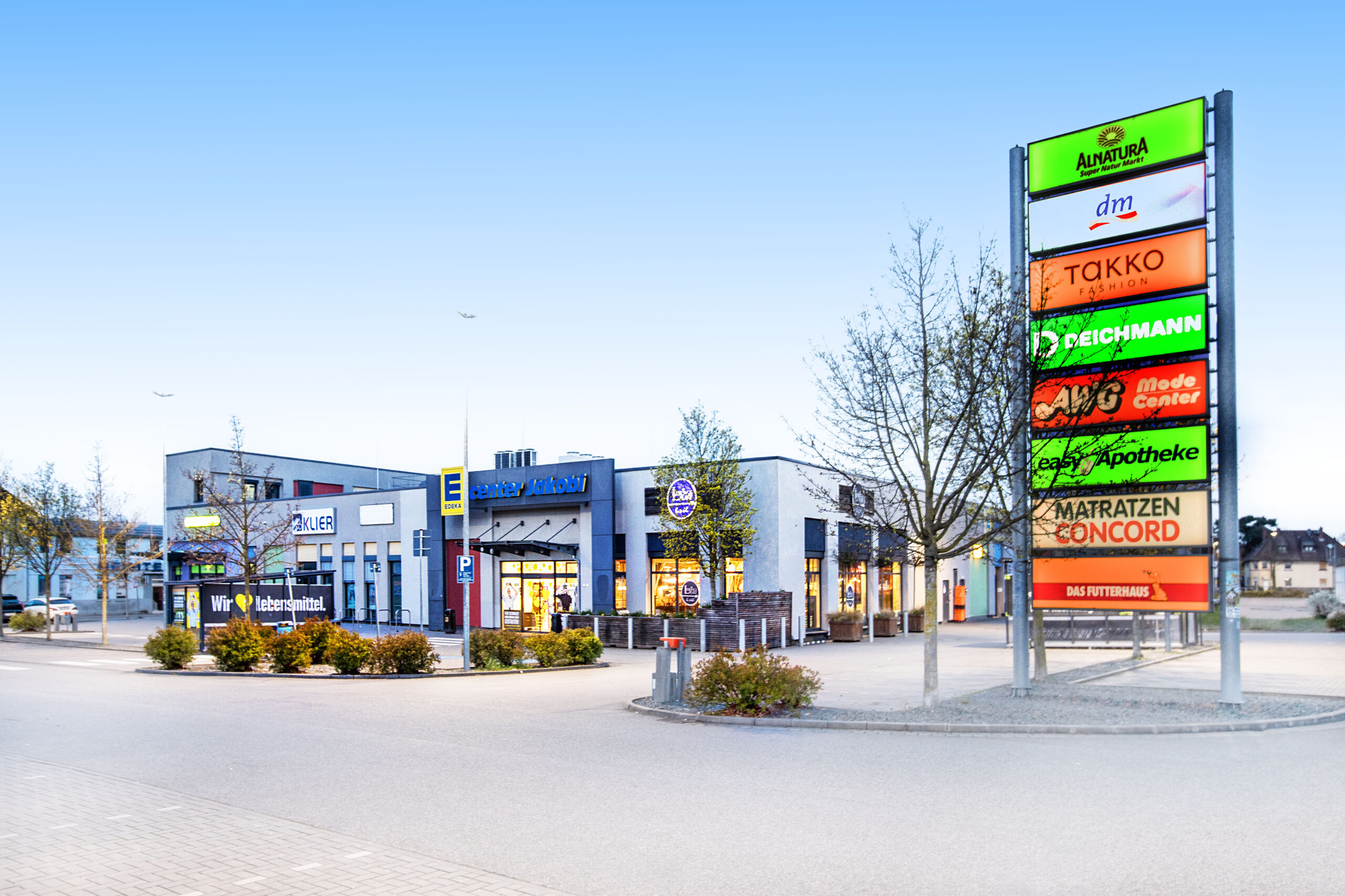Vor einem REWE-Center-Supermarkt mit roten Eingangsschildern stehen Menschen fahren Autos.