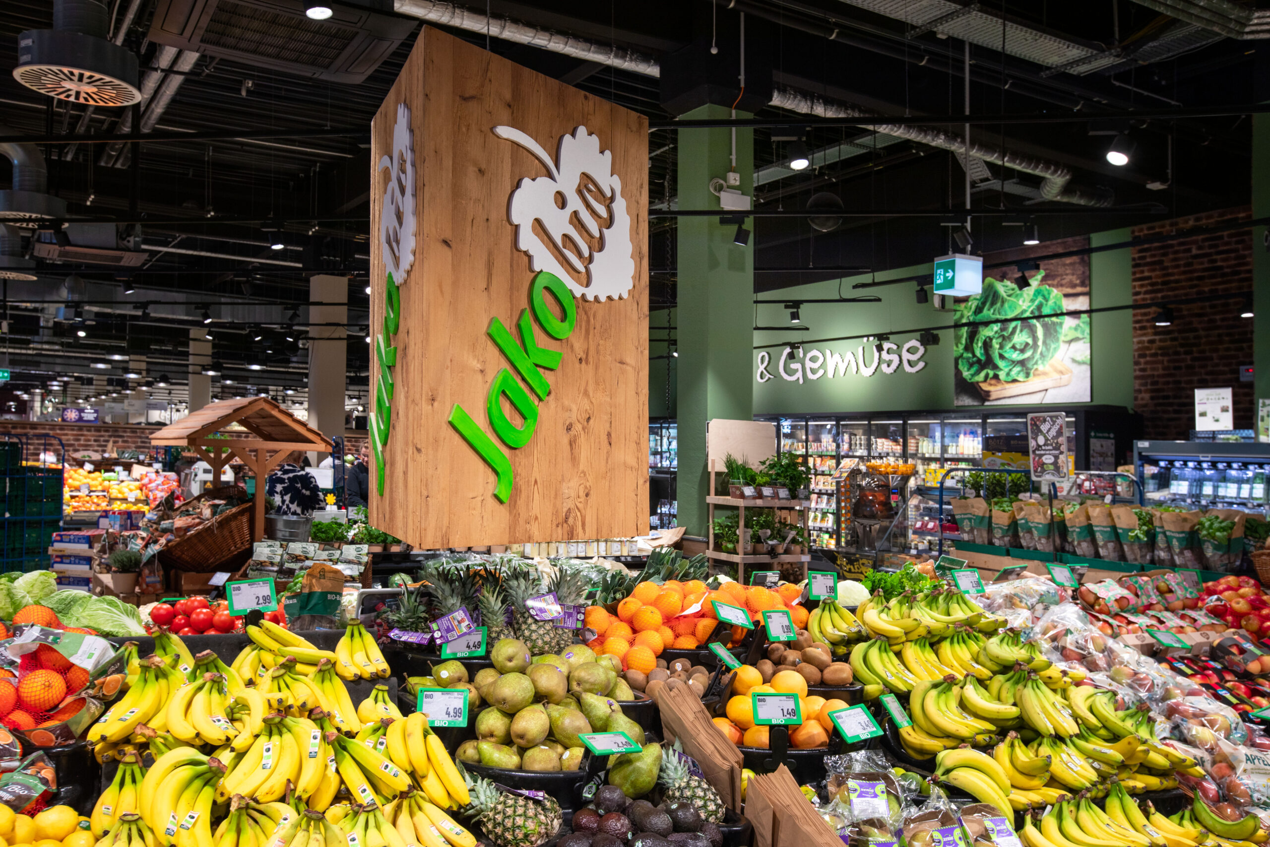 Vor einem REWE-Center-Supermarkt mit roten Eingangsschildern stehen Menschen fahren Autos.